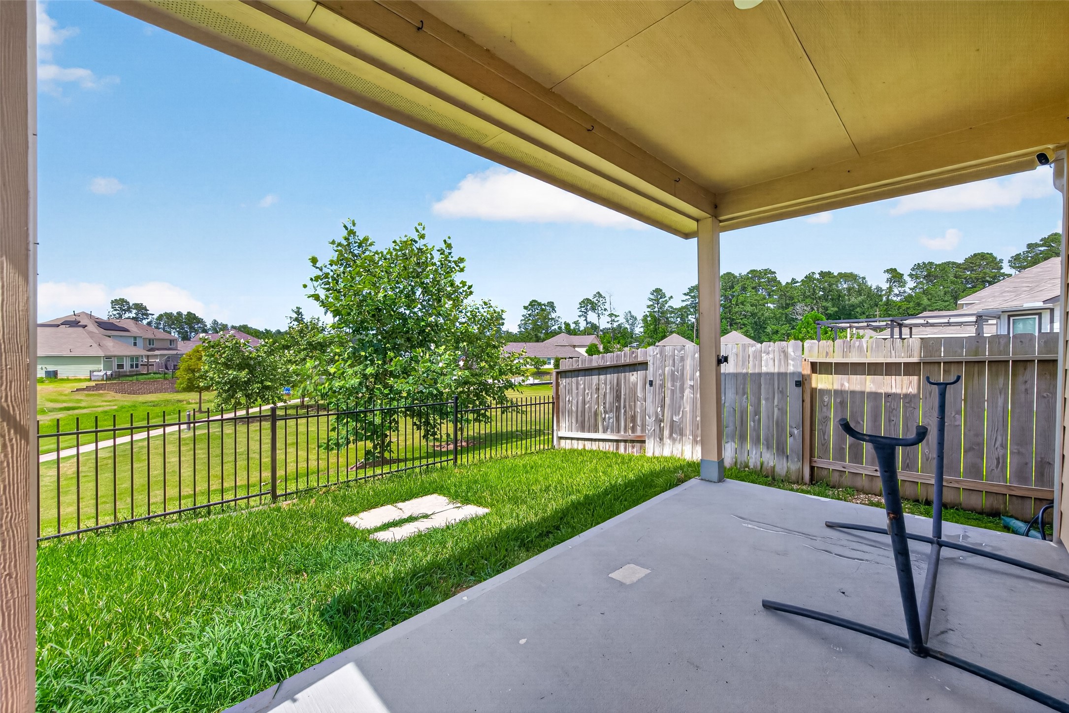 2526 Sturgeon Way Conroe, TX 77384 - Photo 36 of 49 This photo shows a covered patio with a concrete floor, overlooking a fenced backyard with green grass and a tree. The yard backs onto a small path and open green space, with neighboring houses in the background. Sorry, hammock not included!