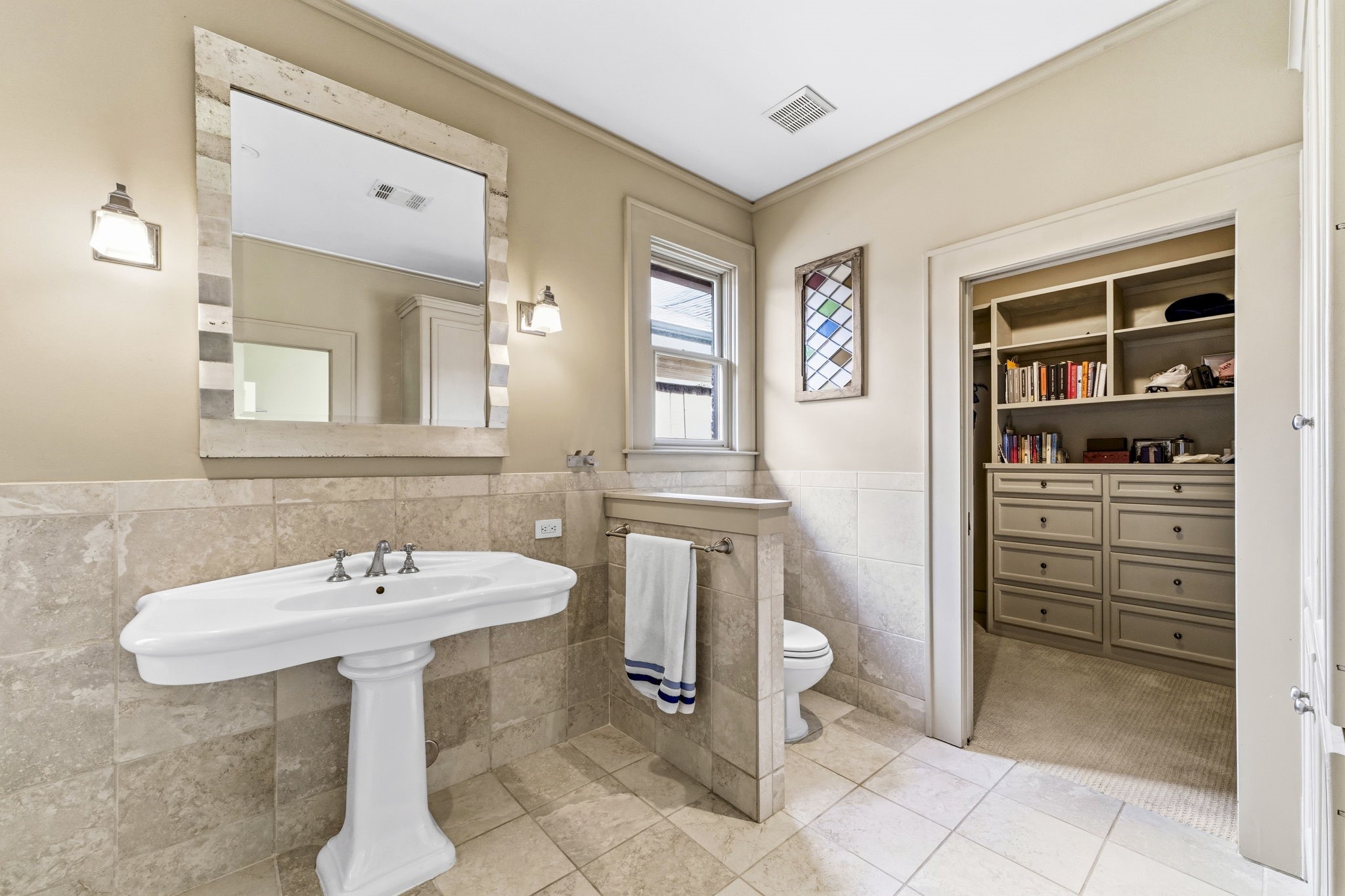 1521 Kipling Street Houston, TX 77006 - Photo 24 of 35 This bathroom features a classic pedestal sink with a large framed mirror, neutral tile flooring, and partial wall tiling. It includes a toilet area with natural light from a window and leads into a spacious walk-in closet with built-in shelves and drawers for ample storage.