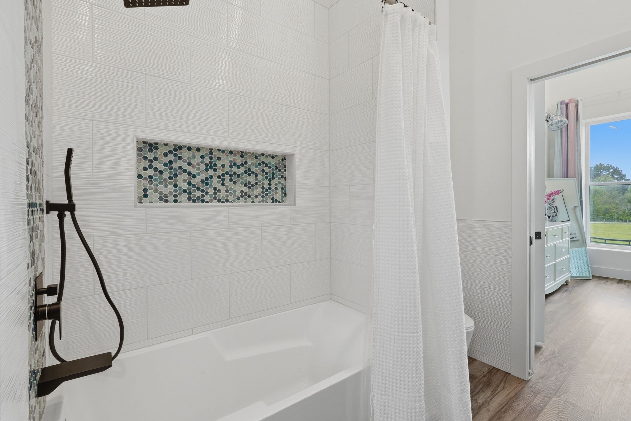 2743 Bobville Road Montgomery, TX 77316 - Photo 36 of 50 This bathroom features a modern tub with a sleek rainfall showerhead and a hexagonal tile accent. A white shower curtain adds elegance, and the room opens to a bright space with wooden flooring.