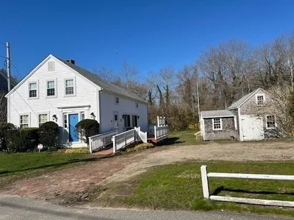 a view of a white house in front of a yard with plants and large tree