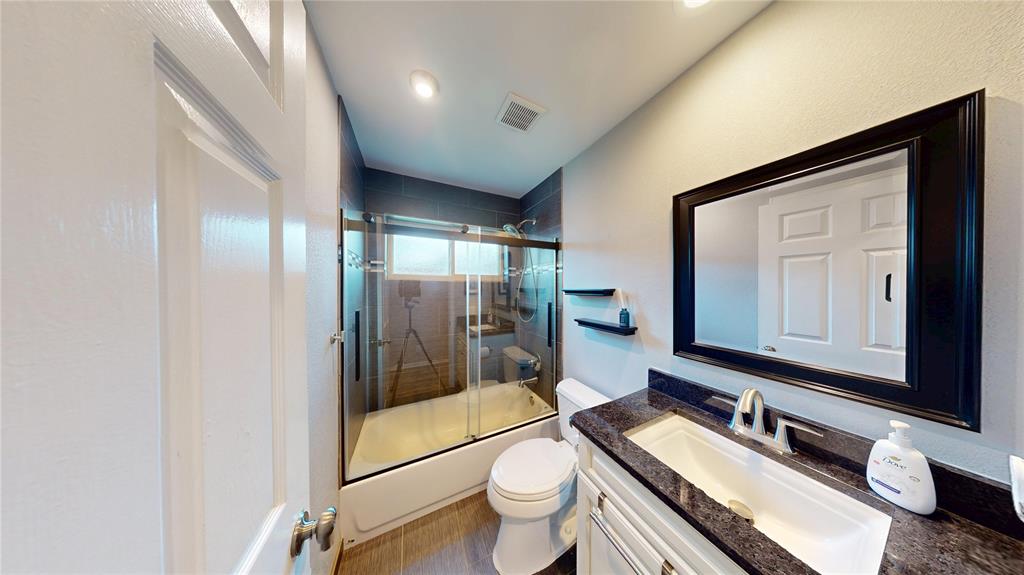 1317 Colonial Place Flower Mound, TX 75028 - Photo 13 of 20 Bathroom featuring a dark tiled shower/tub combination with a glass enclosure, a white vanity with a dark countertop, a rectangular mirror with a dark frame, and wood-finish flooring