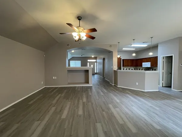 an empty room with wooden floor a ceiling fan and kitchen view