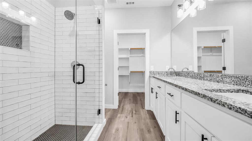 716 East Chestnut Street Denison, TX 75021 - Photo 20 of 26 Spacious bathroom featuring a frameless glass shower enclosure with subway tile surround and mosaic tile floor, dual vanity with granite countertop and undermount sinks, wood-finish flooring, and two open doorways revealing walk-in closets with built-in shelving
