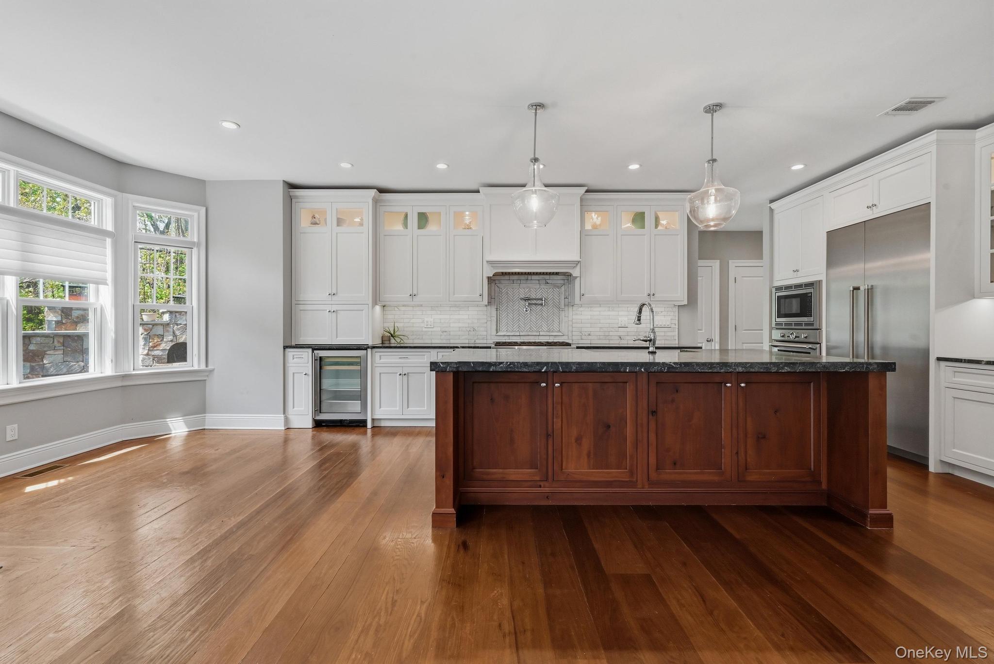 42 Skunks Misery Road Locust Valley, NY 11560 - Photo 11 of 50 Expansive kitchen and dining area featuring wide plank wood-finish flooring, a central island with wood cabinetry and dark countertops, white perimeter cabinetry with integrated appliances, and a bay window