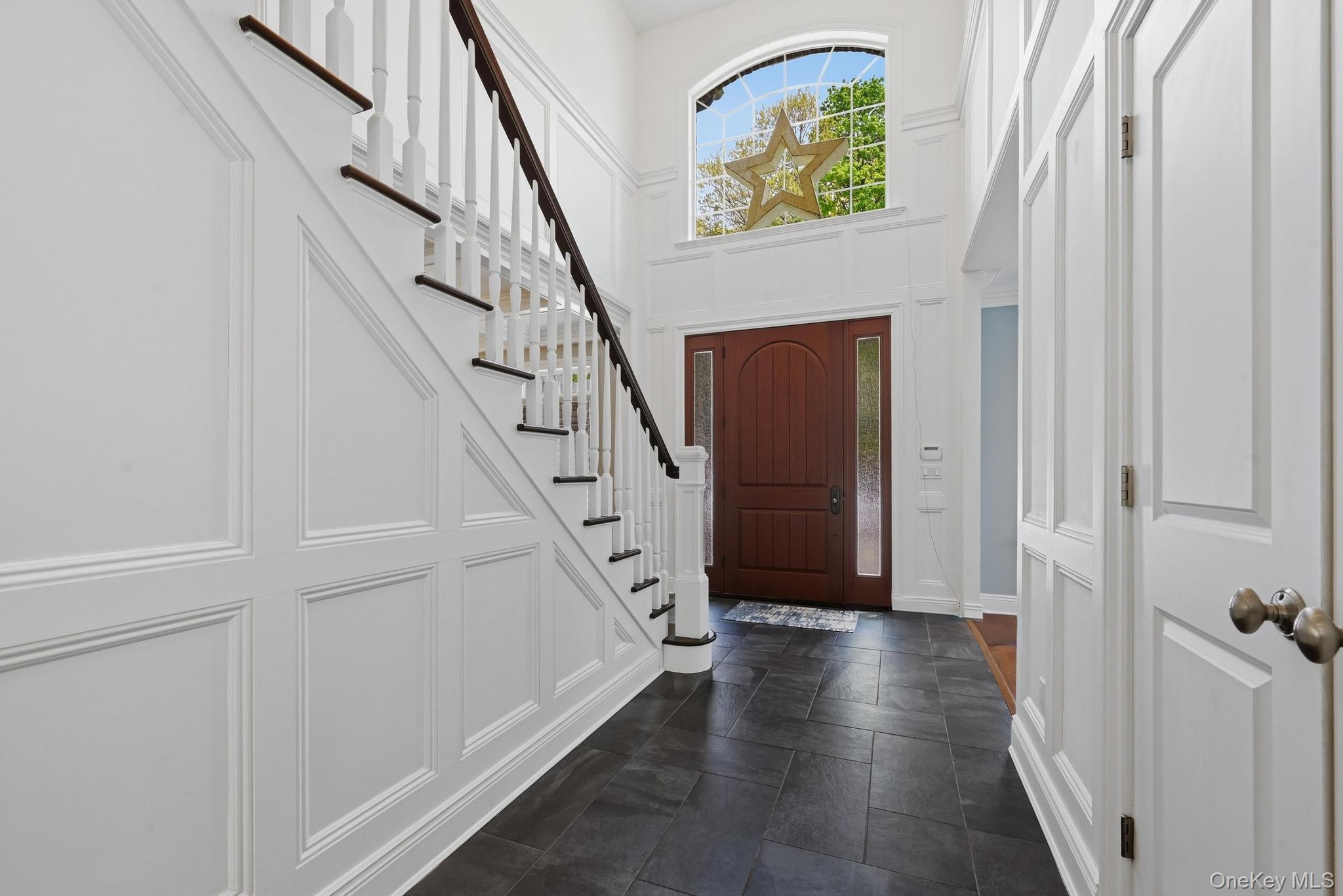 42 Skunks Misery Road Locust Valley, NY 11560 - Photo 2 of 50 Two-story foyer featuring a dark wood entry door with sidelights, arched transom window, extensive white wainscoting, a staircase with dark wood treads and white balusters, and dark tile flooring
