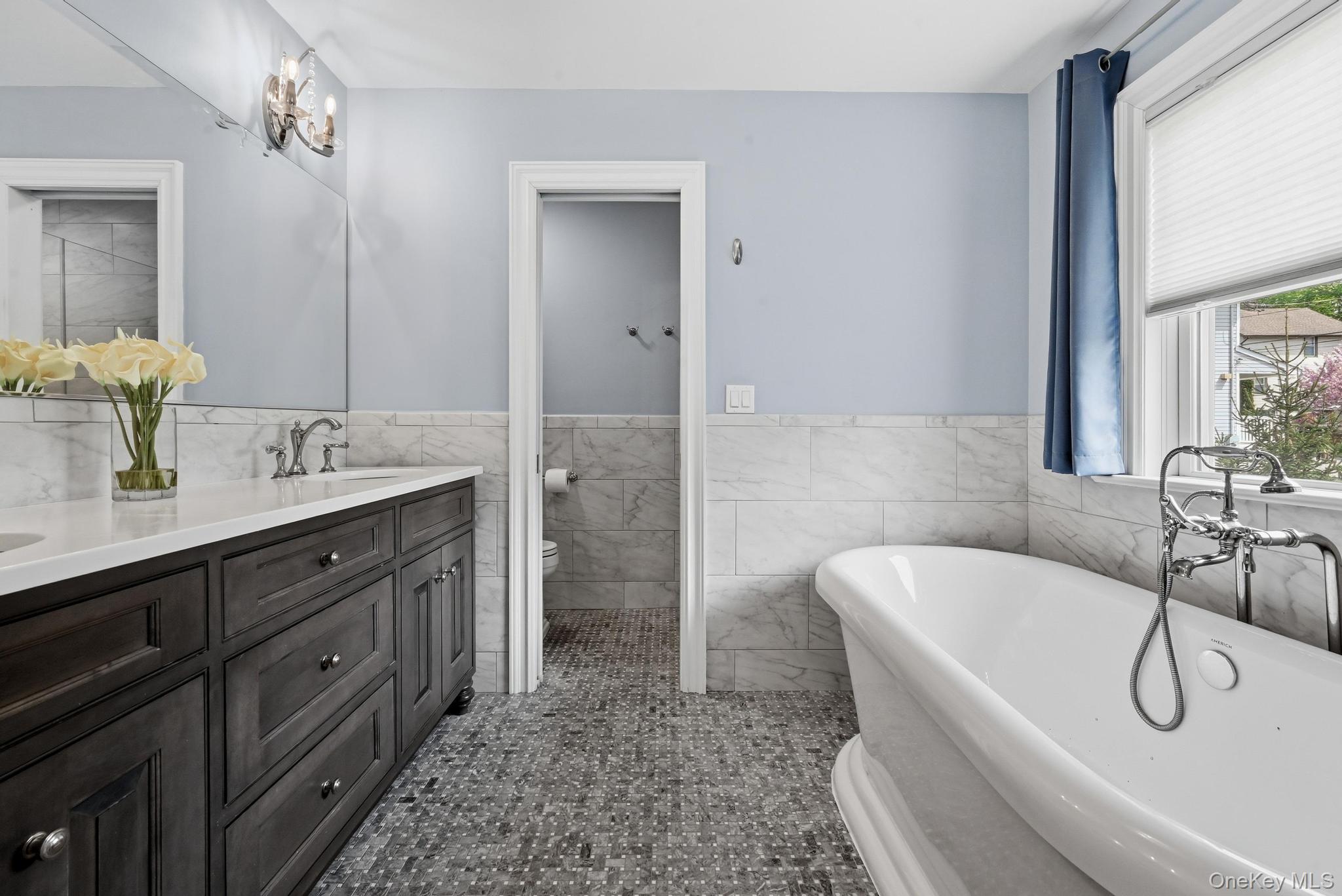 42 Skunks Misery Road Locust Valley, NY 11560 - Photo 27 of 50 Primary Bathroom featuring separate room, freestanding soaking tub with chrome fixtures, dual-sink vanity with dark wood cabinetry and white countertops, large wall-mounted mirror, and stone tile flooring