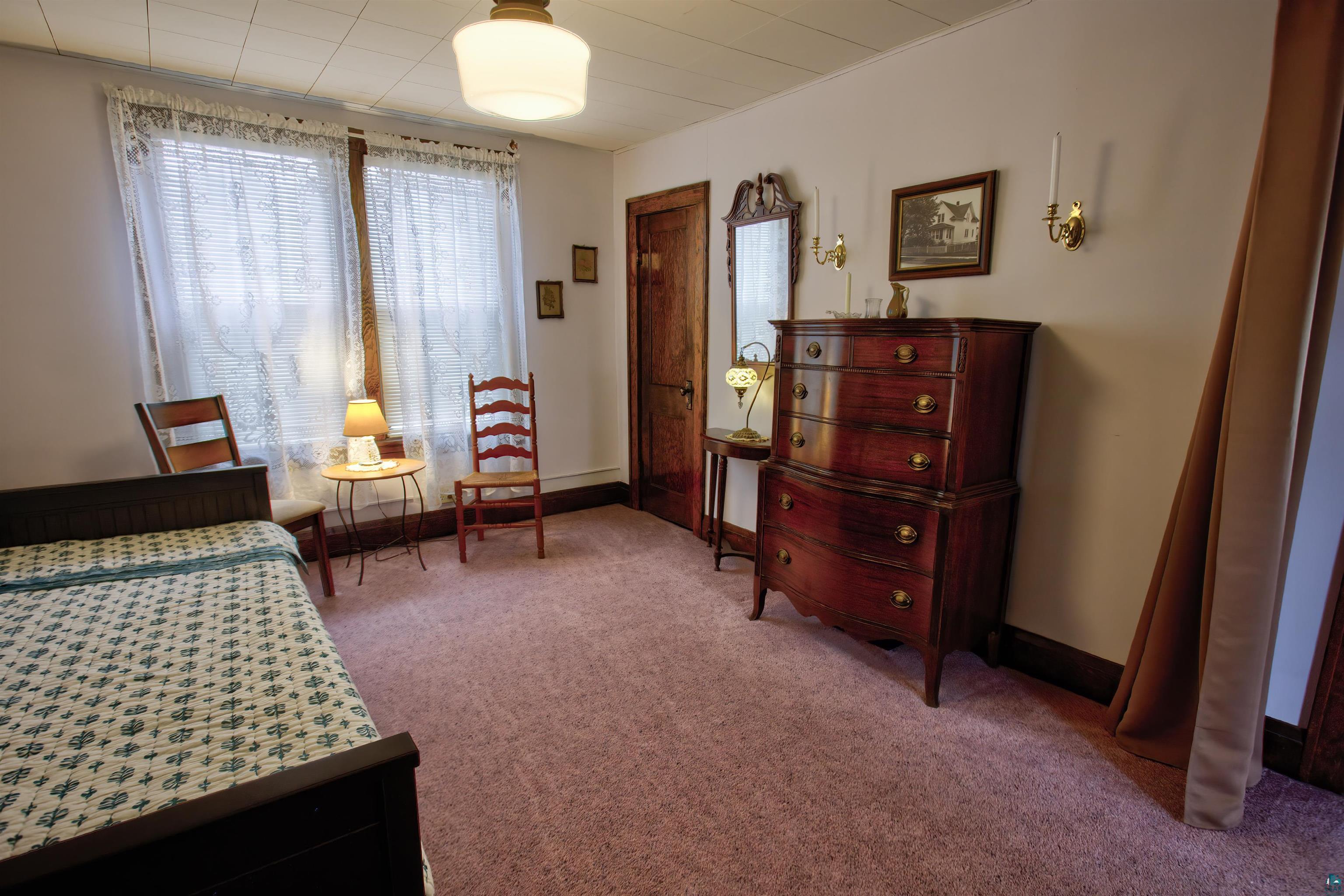 24 East Conan Street Ely, MN 55731 - Photo 15 of 27 Carpeted room featuring dual windows with sheer coverings, dark wood trim, a paneled ceiling, and wall-mounted brass sconces
