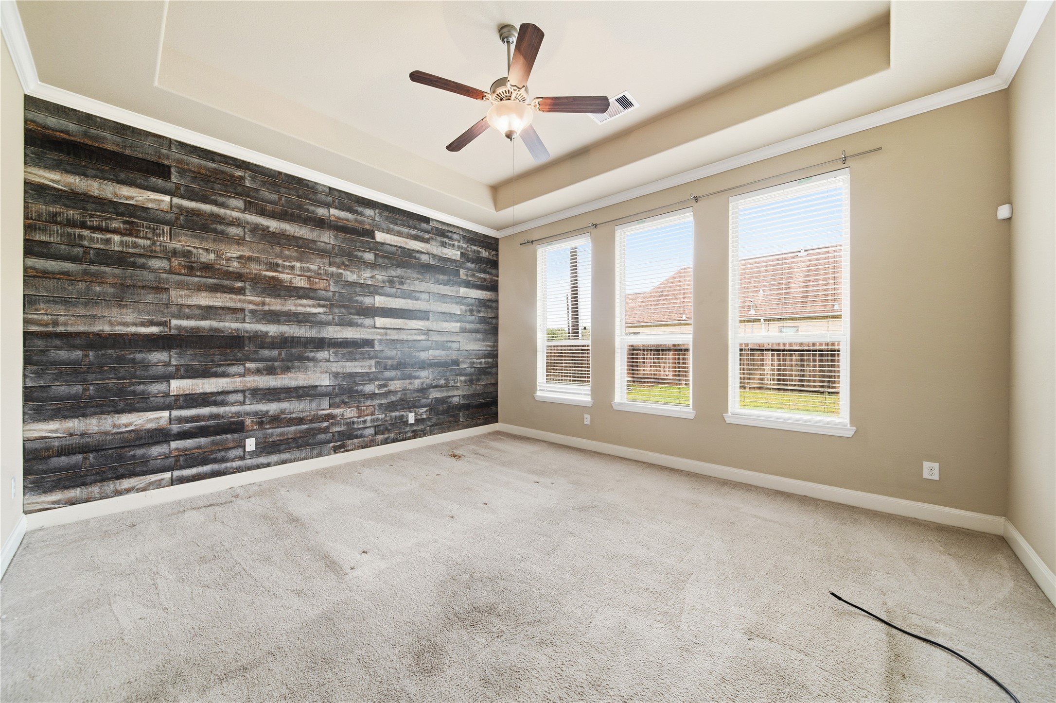 5303 Mesquite Willow Lane Rosharon, TX 77583 - Photo 17 of 32 This primary bedroom features a modern accent wall with dark wood paneling, three large windows offering ample natural light, a ceiling fan, and neutral carpet, creating a bright and versatile space.