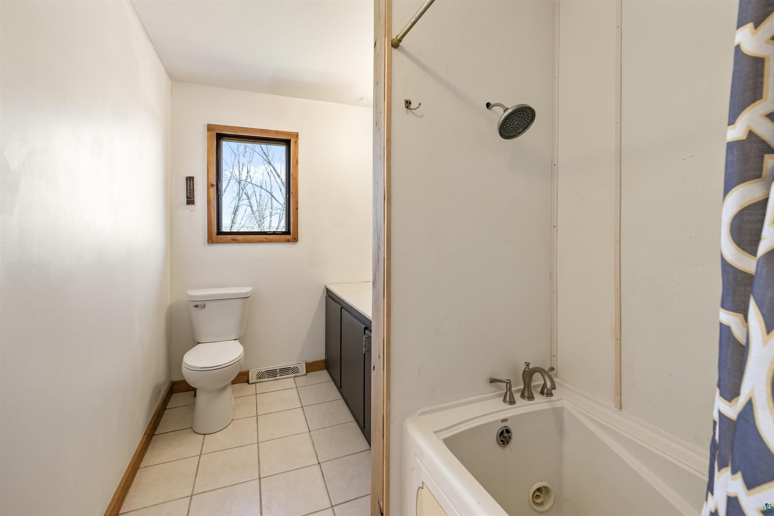 40139 County Road 251 Cohasset, MN 55721 - Photo 22 of 50 Bathroom featuring a window with natural wood trim, white ceramic tile flooring, a white toilet, and a white jetted bathtub with a shower head