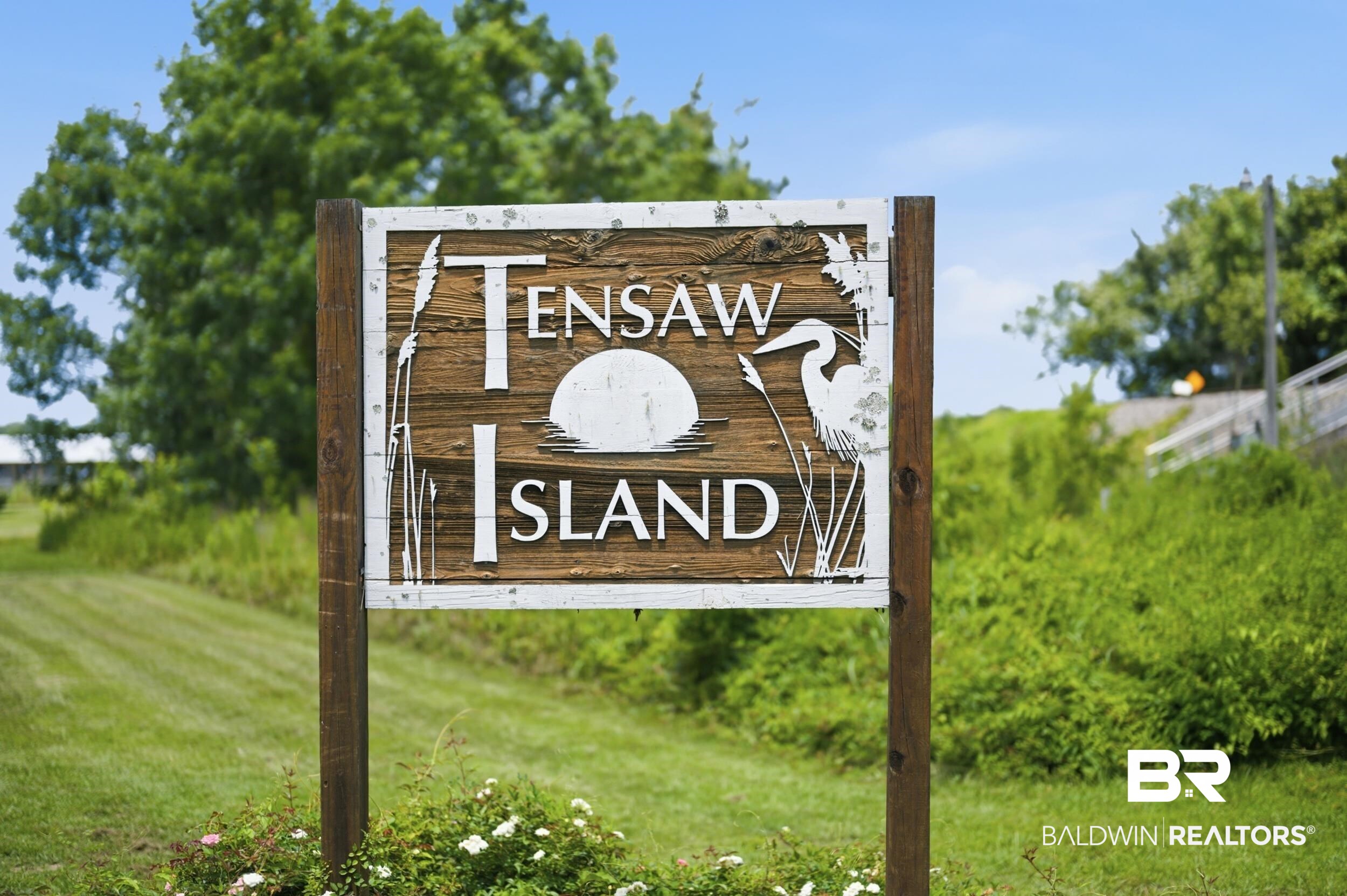 42080 Bayou Road Bay Minette, AL 36507 - Photo 28 of 43 Wooden Tensaw Island sign with white lettering and a heron motif, set against a backdrop of manicured grass, lush green foliage, and mature trees under a clear blue sky