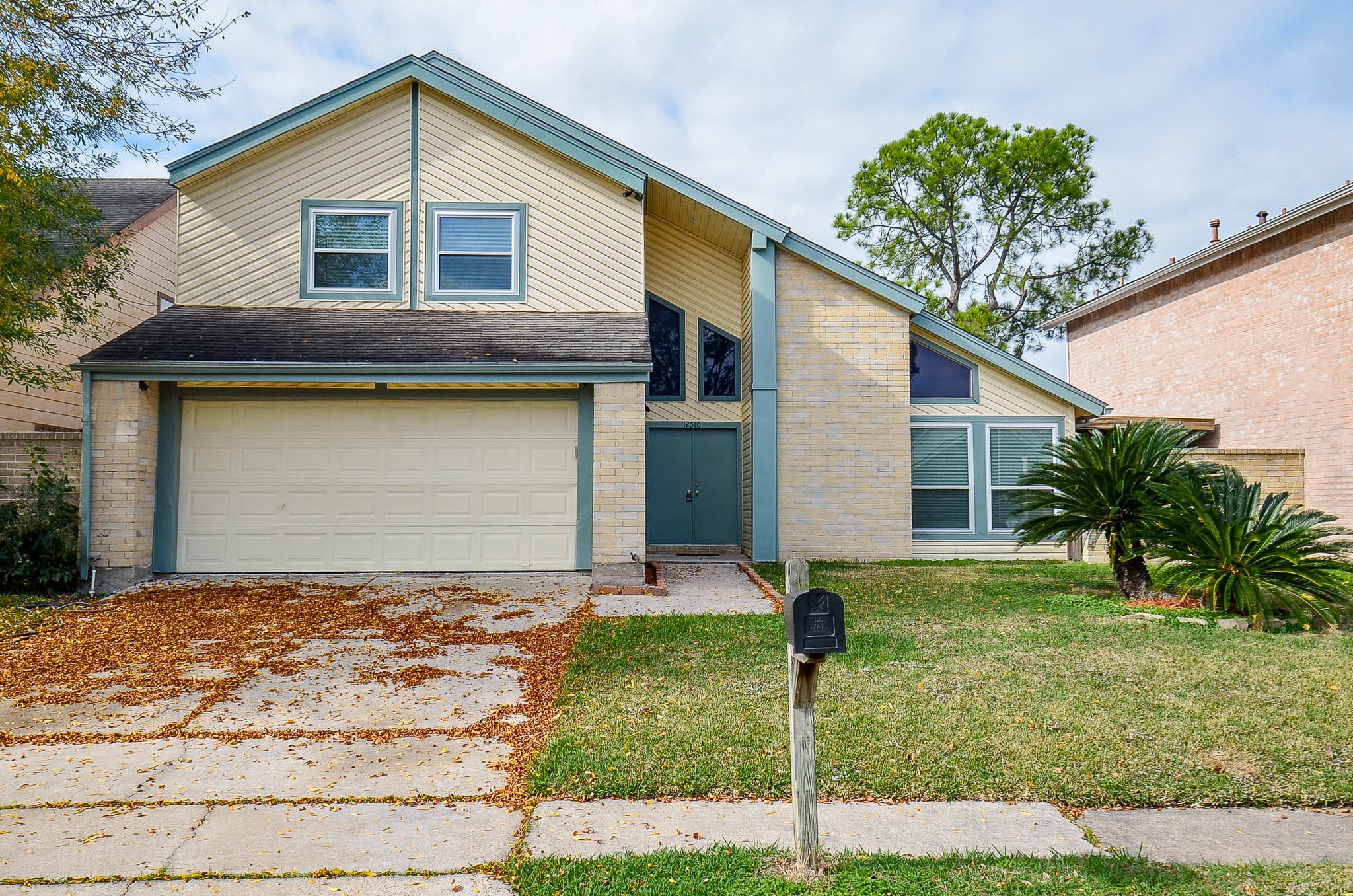 This is a two-story home with a contemporary design featuring a two-car garage and a sloped roof. The exterior is a mix of light brick and siding, with large windows on the second floor. It includes a small front yard with mature trees and shrubs, set in a suburban neighborhood.