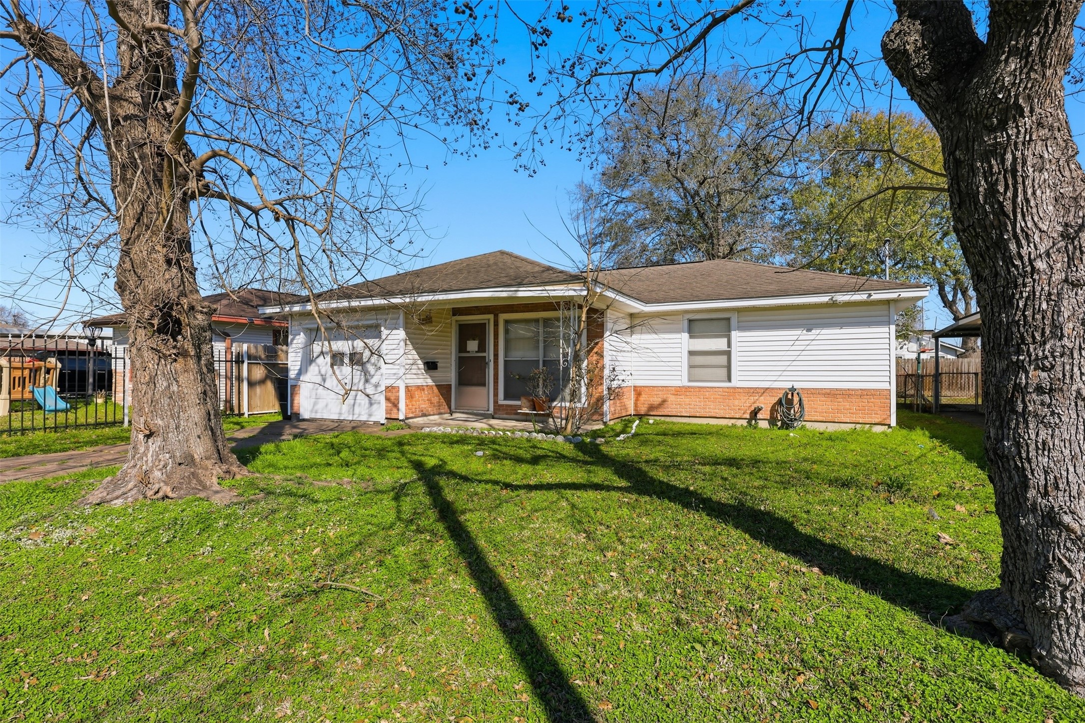 Charming single-story home with a mix of brick and siding exterior, nestled in a lush, grassy yard with mature trees. It features a welcoming front porch and a fenced backyard, perfect for outdoor activities and relaxation.