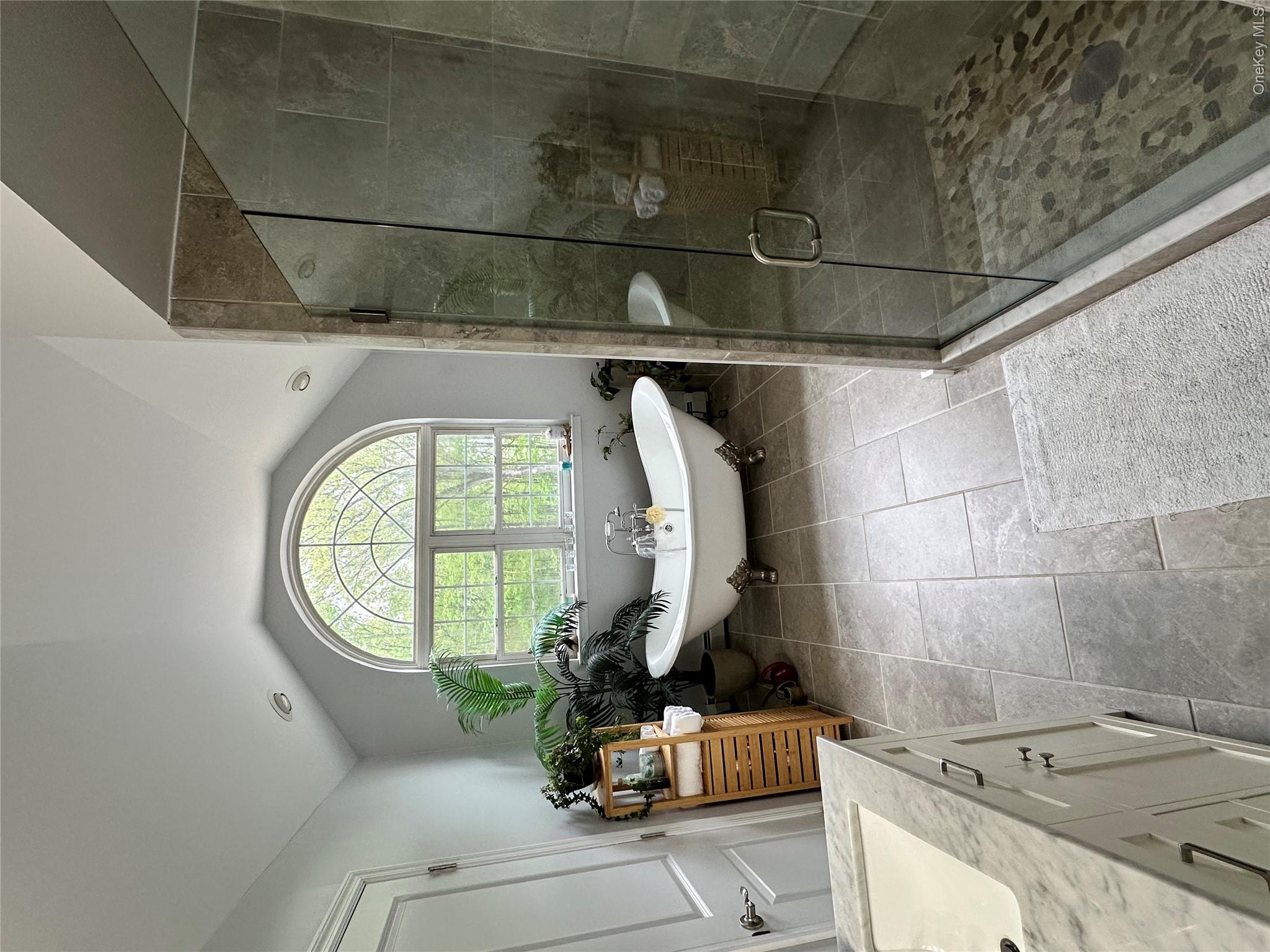 95 Weed Road Pine Bush, NY 12566 - Photo 13 of 31 Spacious bathroom featuring a freestanding clawfoot tub, an arched window, a glass-enclosed shower with pebble flooring, a vanity with an integrated sink and stone countertop, and large format floor tiling