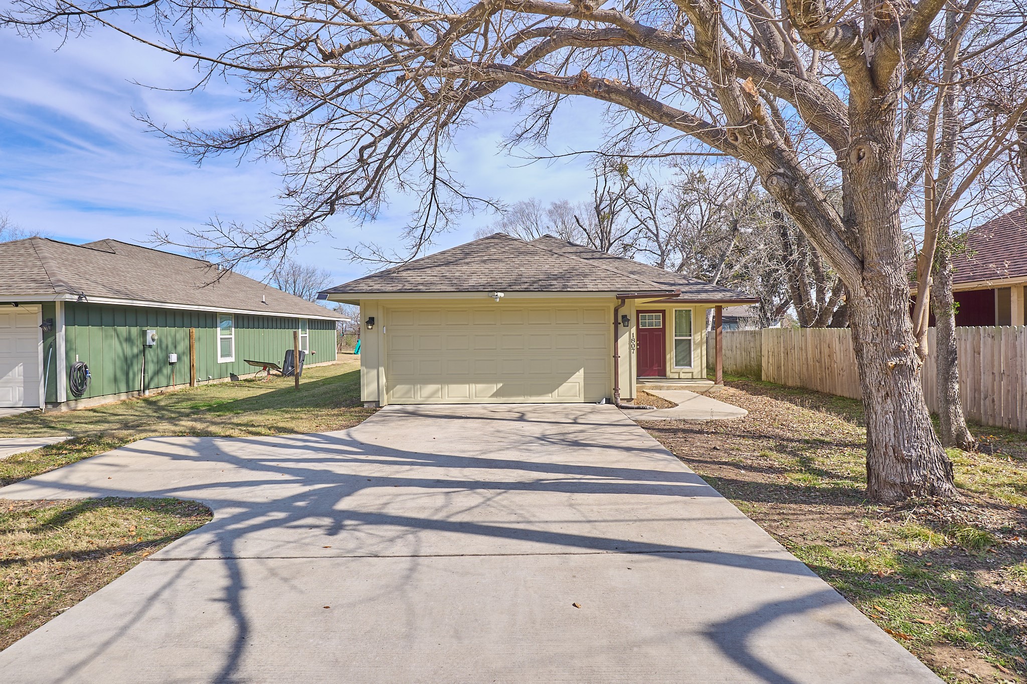 1807 Nuches Lane Bryan, TX 77803 - Photo 3 of 27 This photo shows a single-story home with a two-car garage, a long driveway, and a welcoming front entrance with a red door. The yard is partially shaded by a large tree, and there's a wooden fence on the right. The house is set in a suburban neighborhood with similar homes nearby.
