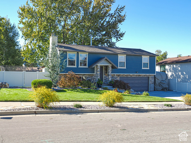 3308 Willow Ridge Road Salt Lake City, UT 84121 - Photo 2 of 59 Split-level residence featuring blue siding, stone facade accents, an attached garage, a white picket fence, and a landscaped front yard with mature trees