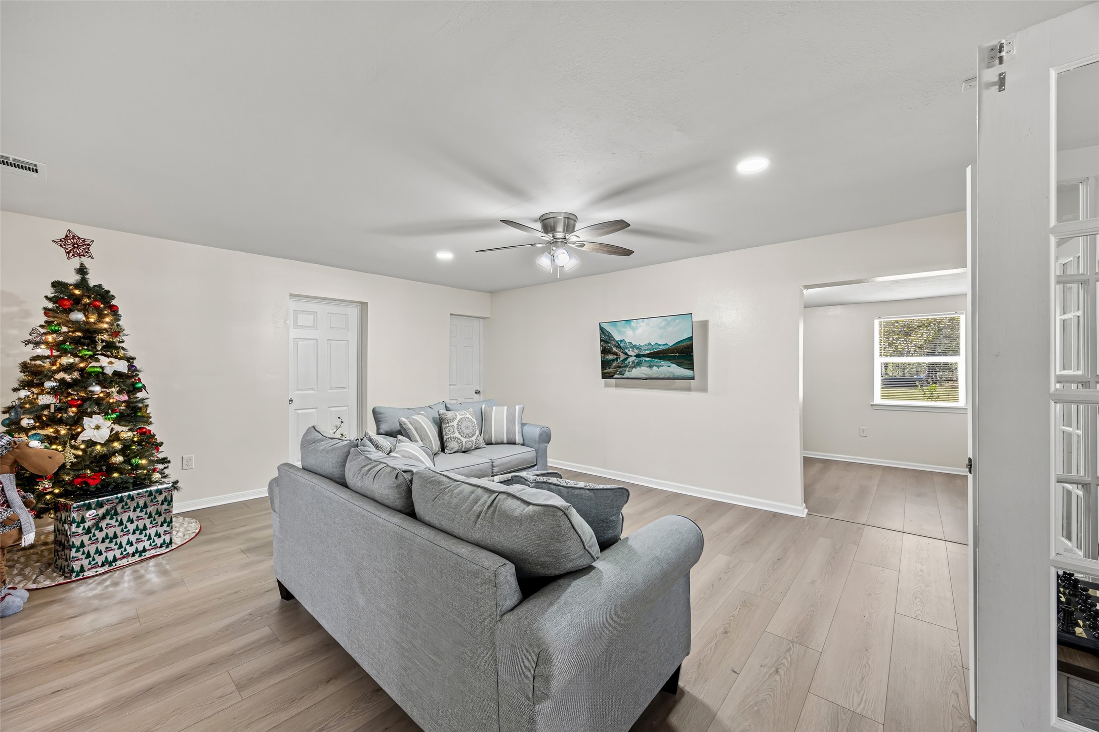 15080 Usher Lane Splendora, TX 77372 - Photo 35 of 44 Spacious living room with modern light wood flooring, neutral walls, and a ceiling fan. Features a cozy gray sofa, Christmas tree, and wall art. Ample natural light from adjacent room's window.