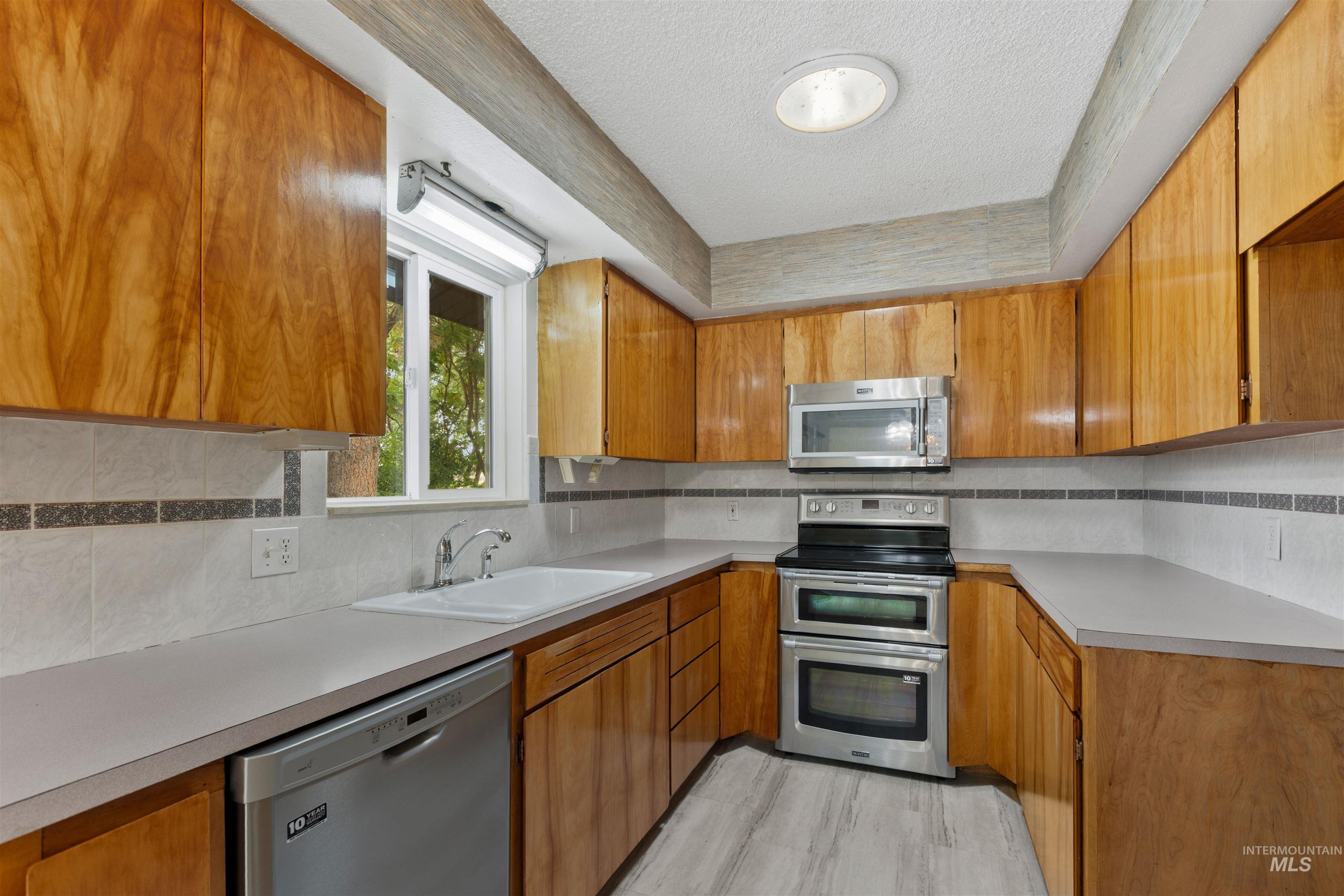 9948 Ash Lane Payette, ID 83661 - Photo 14 of 34 Kitchen with stainless steel appliances, tasteful backsplash, brown cabinetry, light countertops, and a textured ceiling