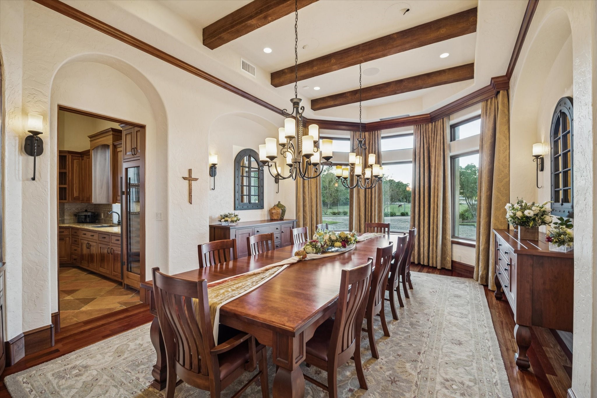 21538 Mueschke Road Tomball, TX 77377 - Photo 13 of 50 Opulent formal dining room. Hosting 10–12 guests, this exquisite space features a tray ceiling with rich wood beams, wrought-iron chandeliers, and a bay of windows with Lutron shades overlooking the courtyard, front grounds, and the adjoining butler’s kitchen. The refined details and luxurious finishes create a sophisticated setting for memorable gatherings.