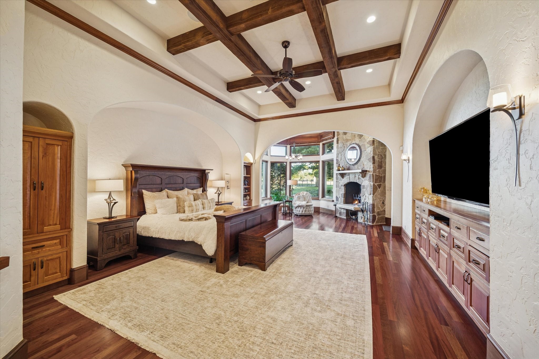 21538 Mueschke Road Tomball, TX 77377 - Photo 17 of 50 Primary bedroom sanctuary. This expansive retreat exudes Mediterranean elegance with graceful arches, wood beams on the tray ceiling, crown moldings, and a stone fireplace. A seating area offers stunning views and direct access to the patio and pool area, along with a majestic pine tree, creating a serene and elevated atmosphere. Situated in the west wing, the suite provides ultimate privacy and unparalleled comfort.
