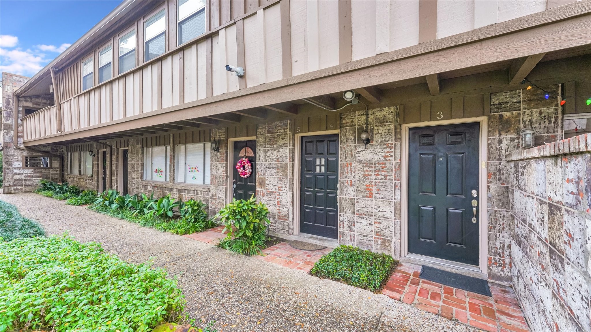 Charming two-story townhouse exterior with a rustic brick facade and dark doors. The landscaped pathway offers a welcoming entrance, complemented by greenery and a cozy ambiance.