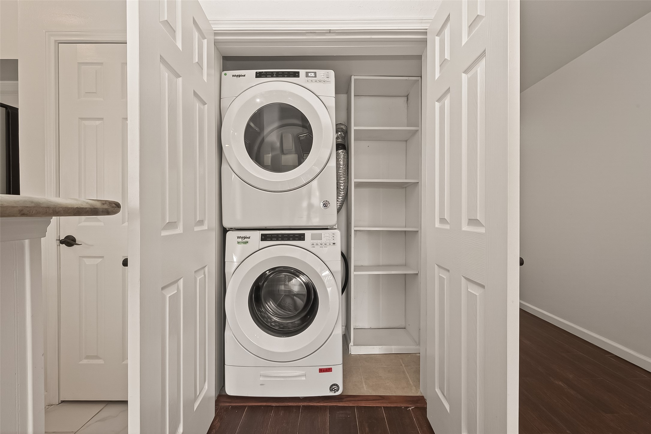180 April Point Drive South Conroe, TX 77356 - Photo 22 of 39 This photo shows a compact laundry area featuring a stacked washer and dryer set within a closet. There are built-in shelves for additional storage on the side, and the space is enclosed by double doors. The flooring is a mix of tile and wood, providing a neat and functional setup.