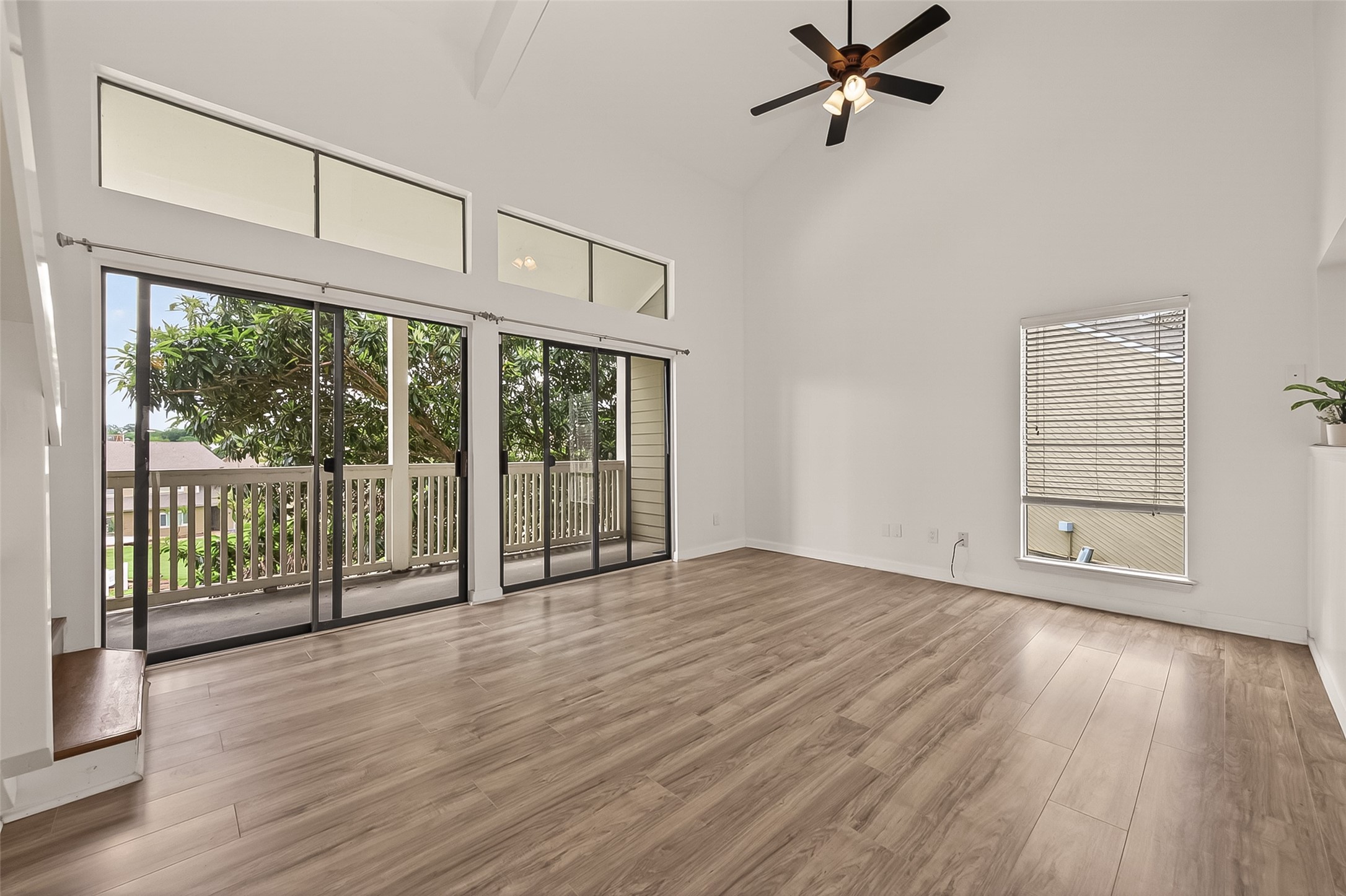 180 April Point Drive South Conroe, TX 77356 - Photo 25 of 39 This spacious living area features high ceilings, large windows for abundant natural light, and sliding glass doors that open to a balcony with a view of greenery. The room is accented by a ceiling fan and has elegant wood flooring.