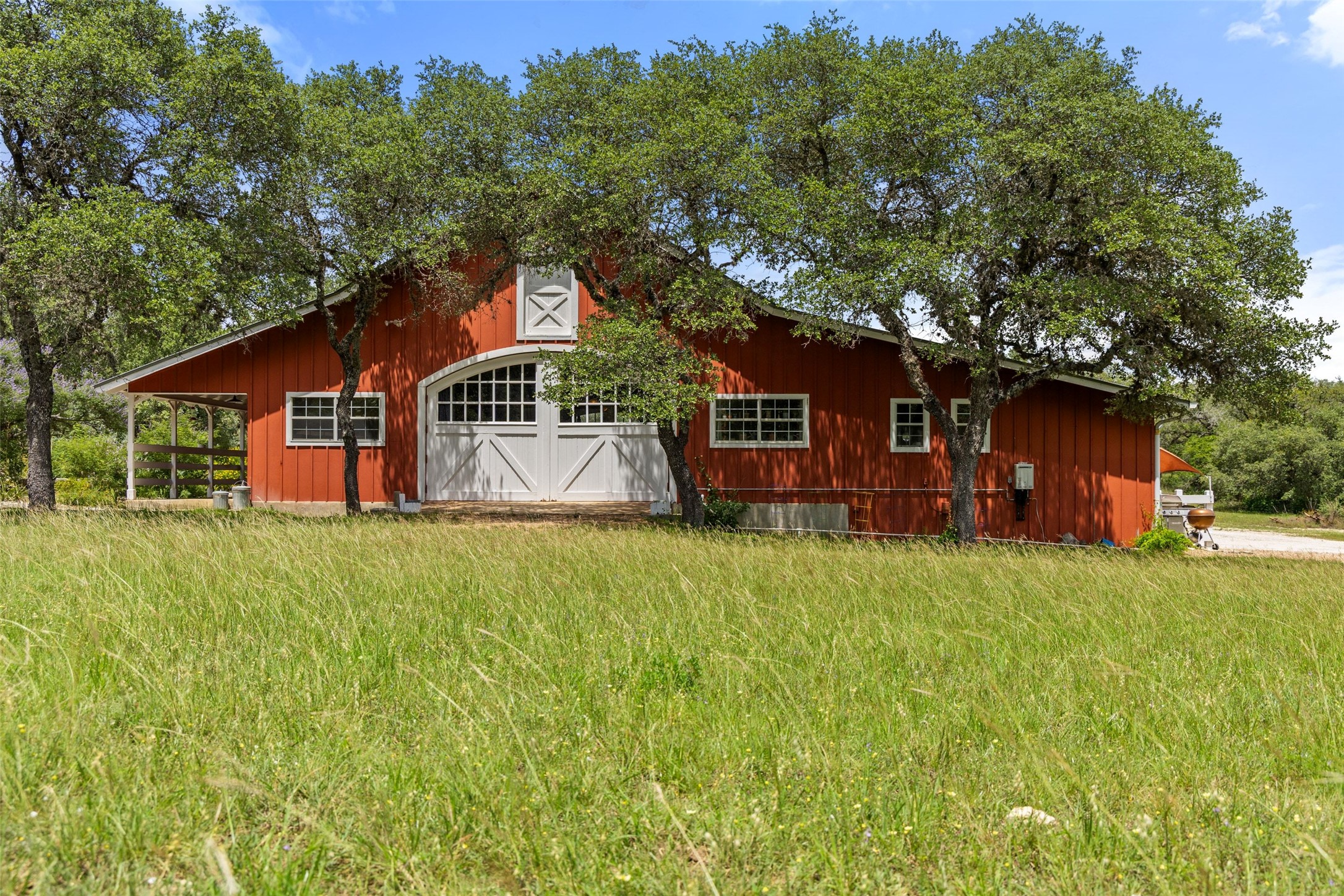 505 Red Corral Ranch Road Wimberley, TX 78676 - Photo 36 of 40 A picturesque red barn nestled among mature trees, showcasing classic white crossbuck doors.
