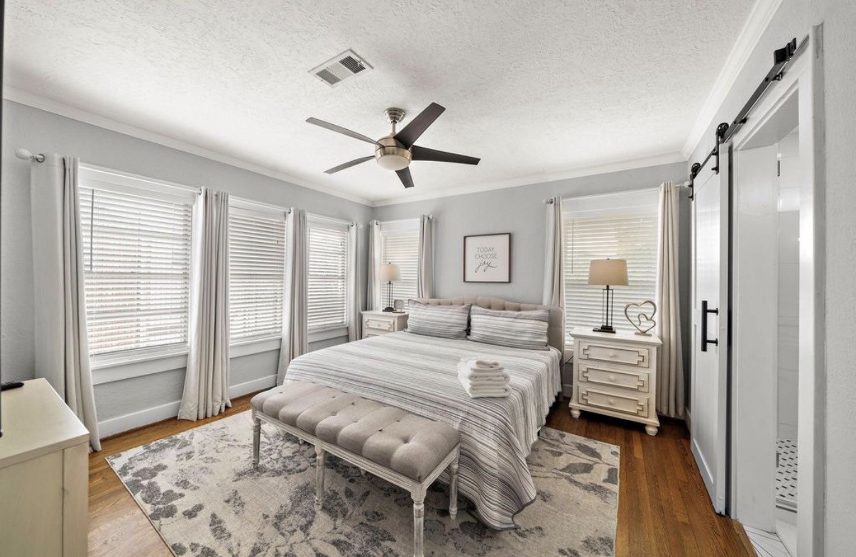 1633 Castle Court Houston, TX 77006 - Photo 14 of 38 Staged primary bedroom showcasing abundant natural light from multiple windows, warm wood flooring, and a serene, neutral palette. The spacious layout includes room for a king bed, seating, and décor, complemented by a stylish barn door leading to the en-suite bath—creating a calm, inviting retreat.