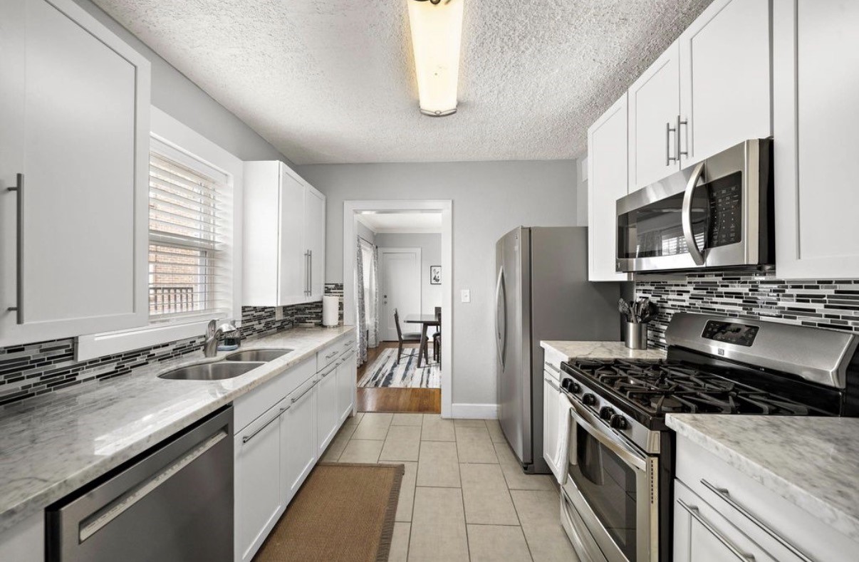 1633 Castle Court Houston, TX 77006 - Photo 4 of 38 Staged modern kitchen featuring sleek white cabinetry, quartz countertops, stainless steel appliances, and a stylish tile backsplash. This bright galley layout offers ample storage, generous counter space, and a seamless flow into the dining area—perfect for everyday cooking and entertaining.