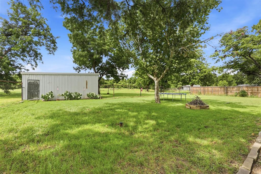 308 West 1st Street Springtown, TX 76082 - Photo 27 of 39 Expansive green space featuring a corrugated metal outbuilding with a single door, established trees, and a wooden privacy fence