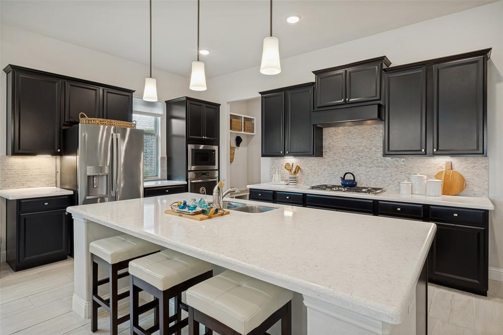 7821 Kentmere The Colony, TX 75056 - Photo 6 of 40 Designed for both everyday living and entertaining, the gourmet kitchen showcases an oversized quartz island with seating, rich cabinetry, stainless steel appliances, a gas cooktop, designer pendant lighting, and a beautiful tile backsplash. A spacious walk-in pantry and open-concept layout complete this stylish, functional space.