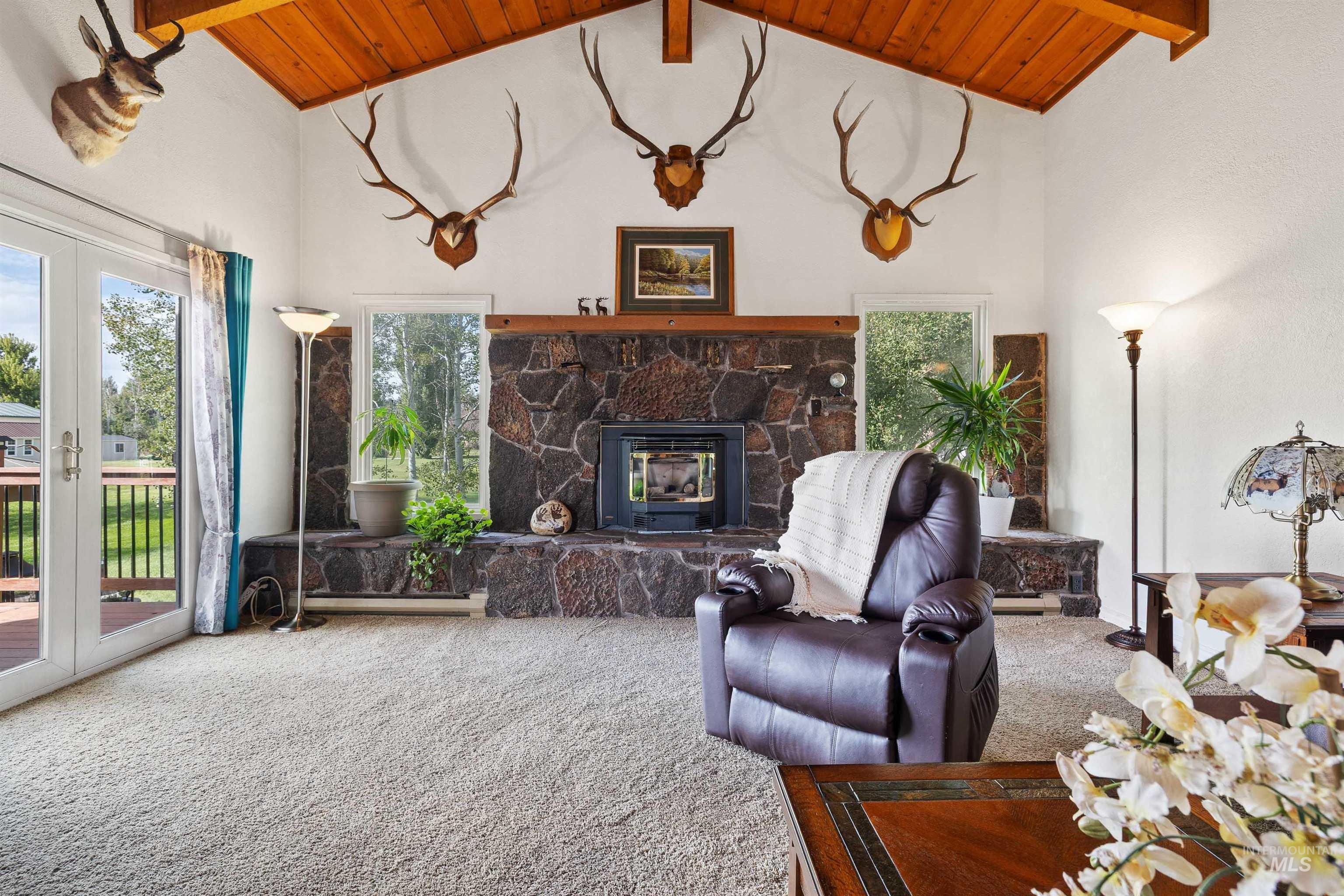 Vaulted wood-paneled ceiling, stone fireplace with hearth, wood mantel, and large picture windows, with double patio doors to an outdoor deck