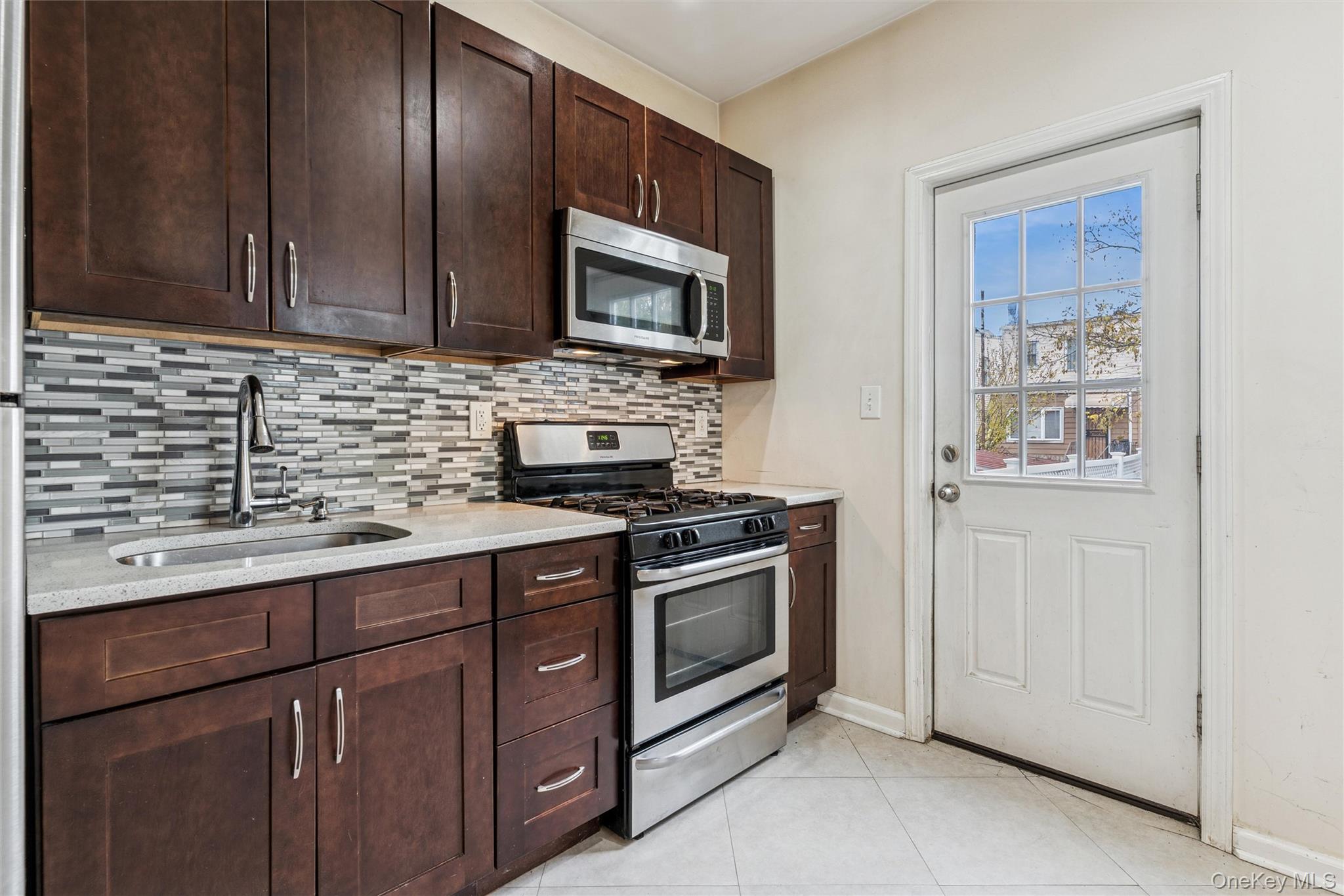 91-25 90th Street Queens, NY 11421 - Photo 12 of 30 Kitchen with stainless steel appliances, dark brown cabinetry, decorative backsplash, light tile patterned floors, and light stone countertops