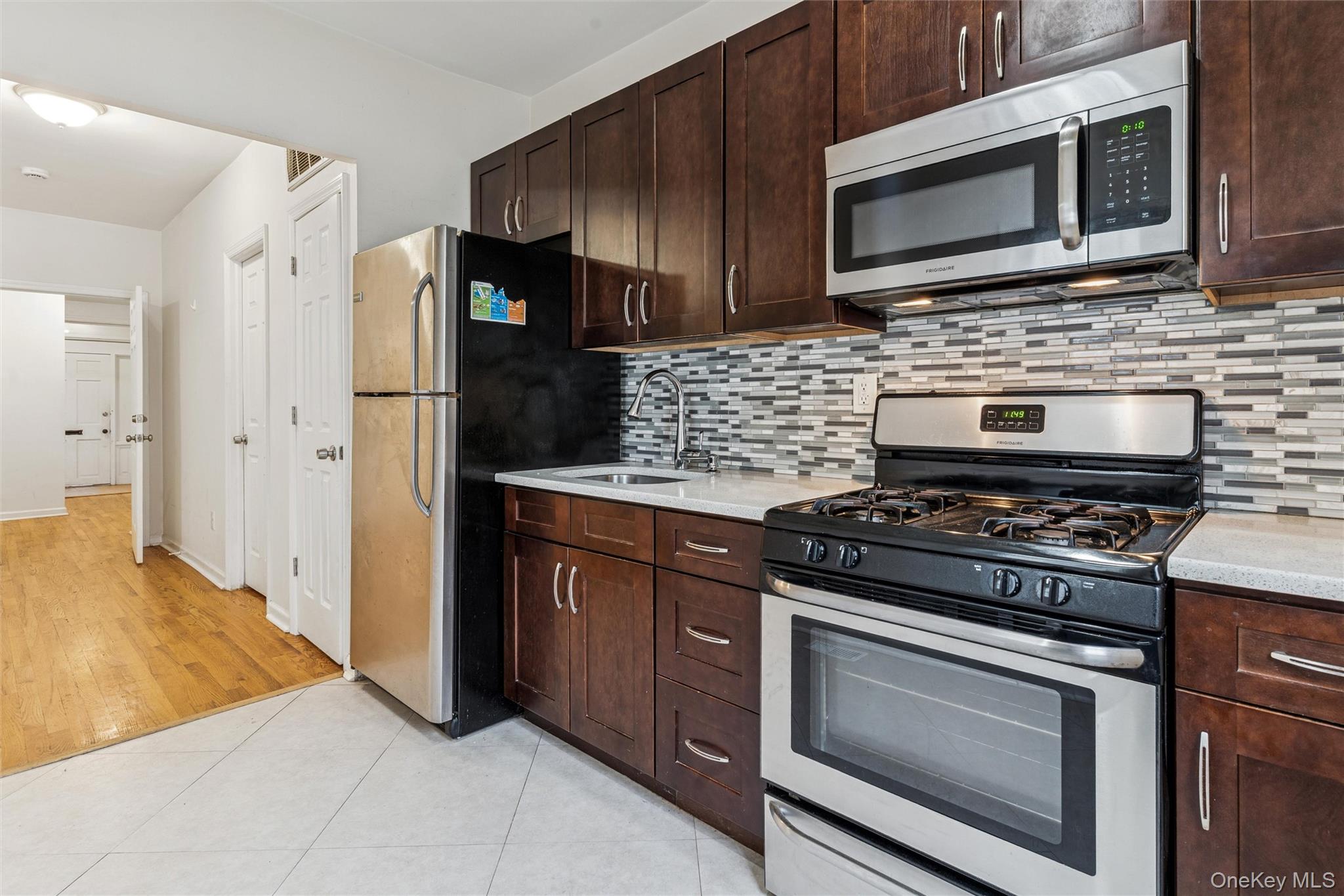 91-25 90th Street Queens, NY 11421 - Photo 13 of 30 Kitchen featuring appliances with stainless steel finishes, backsplash, light tile patterned flooring, dark brown cabinets, and light stone countertops