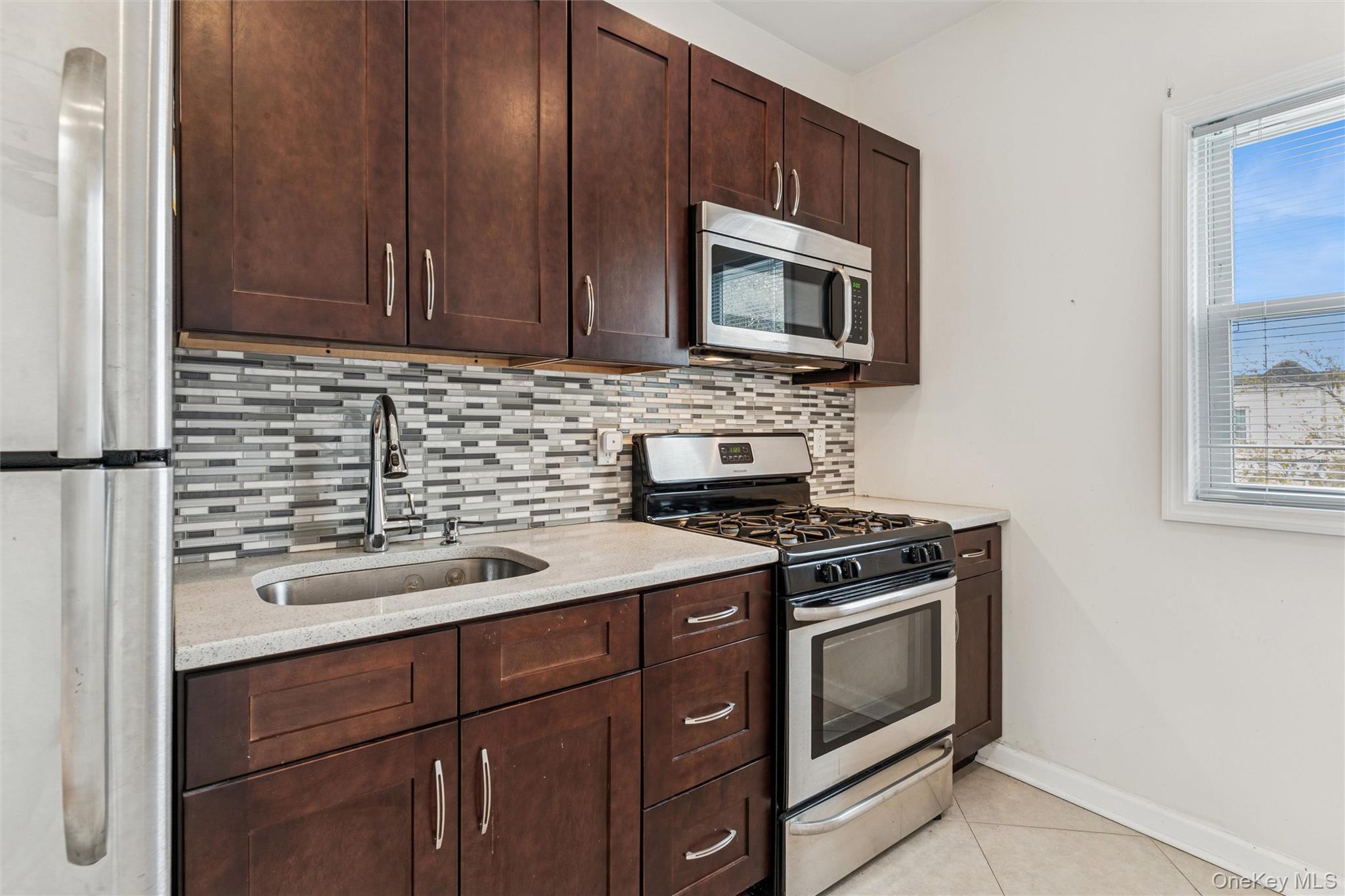 91-25 90th Street Queens, NY 11421 - Photo 20 of 30 Kitchen with appliances with stainless steel finishes, backsplash, light tile patterned floors, dark brown cabinets, and light stone counters