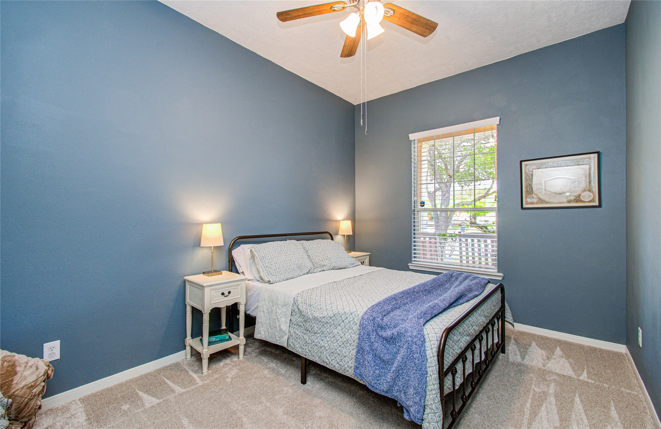 20007 Alcea Court Spring, TX 77379 - Photo 24 of 36 This cozy bedroom features soft blue walls, a ceiling fan, and a large window allowing natural light. It includes a comfortable bed with side tables and lamps, offering a warm and inviting atmosphere.