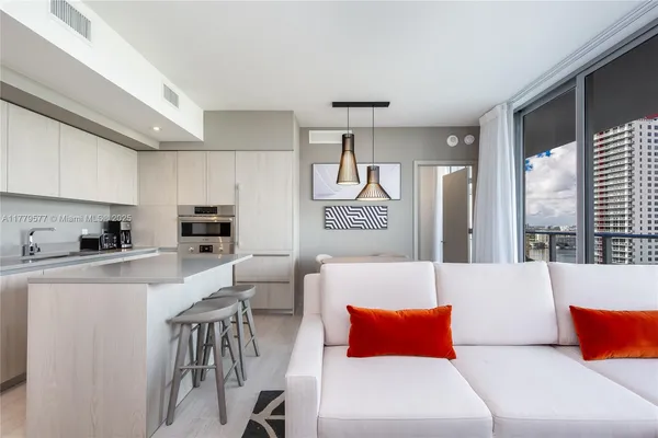 a living room with stainless steel appliances furniture a large window and a kitchen view