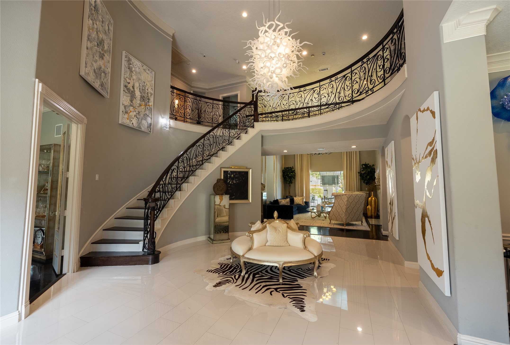 4002 Silver Ridge Boulevard Missouri City, TX 77459 - Photo 5 of 50 This is a spacious and elegantly designed entryway featuring a grand curved staircase with intricate wrought-iron railings, polished tile flooring, and a modern chandelier. The area is well-lit and opens into a comfortable living space, complete with tasteful artwork and sophisticated decor.