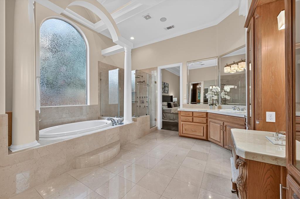 6416 Riverside Drive Plano, TX 75024 - Photo 17 of 35 Spacious bathroom featuring a built-in tub, arched privacy window, decorative columns, a glass-enclosed shower, and dual vanity areas with wood cabinetry and light-toned countertops