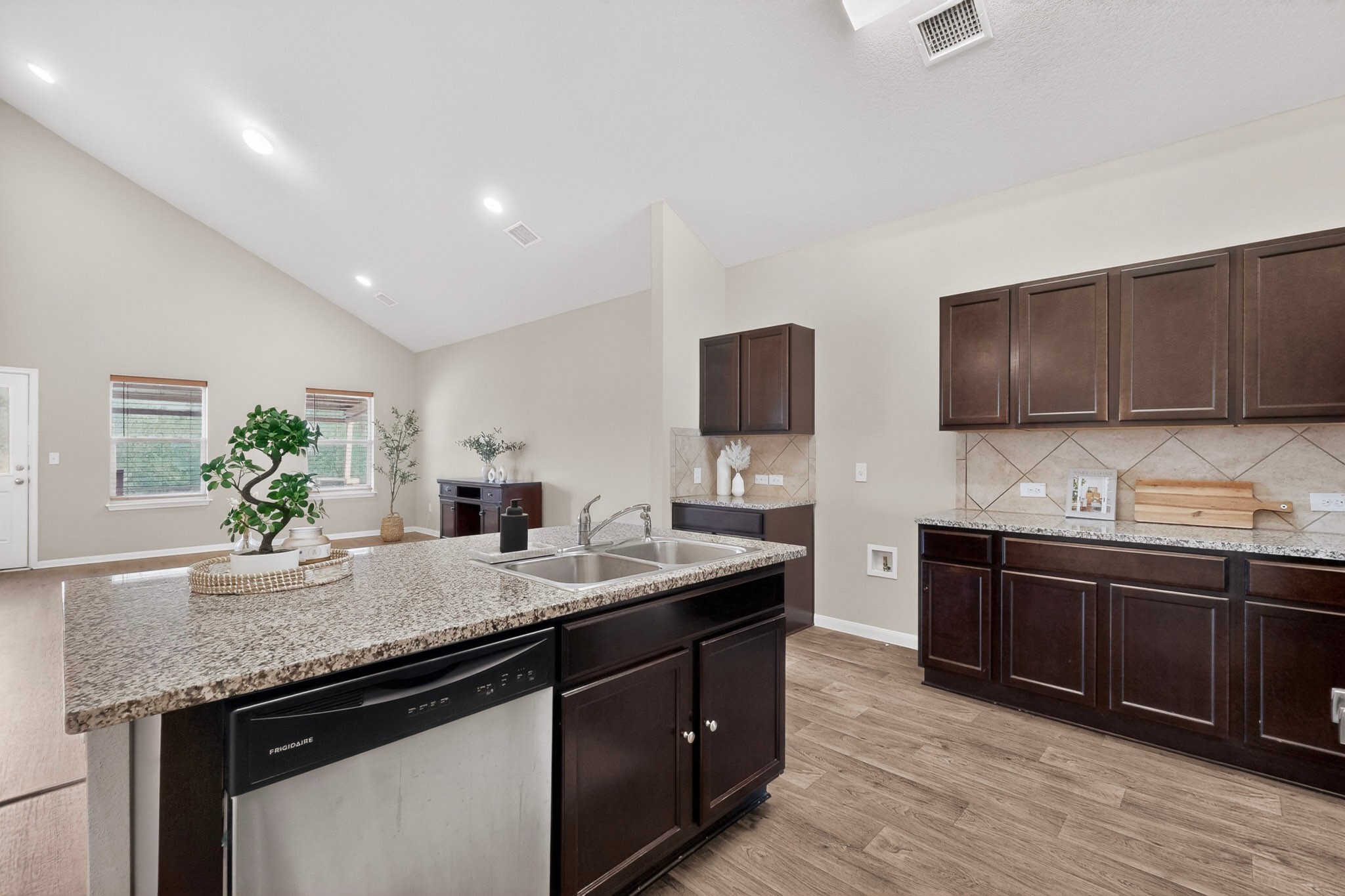 5905 Diamondleaf Bend Austin, TX 78724 - Photo 14 of 38 Open-concept kitchen featuring an island with a double basin sink and stainless steel dishwasher, wood-finish flooring, dark wood cabinetry, and light-toned countertops with a tile backsplash