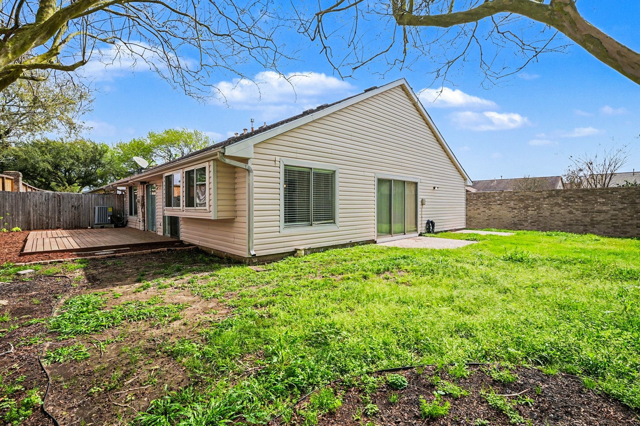 1723 Linfield Way Houston, TX 77058 - Photo 21 of 21 This photo showcases a spacious backyard featuring a single-story home with light siding, large windows, and a sliding glass door. The yard includes a wooden deck area and is bordered by a wooden fence and brick wall, offering privacy and ample outdoor space.