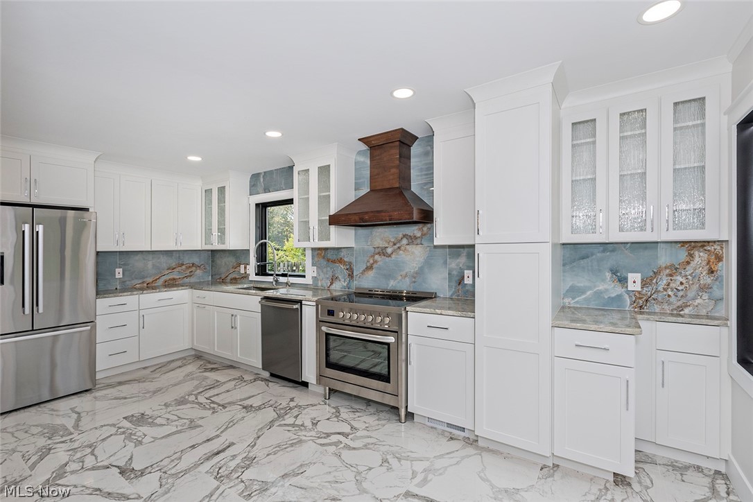 20601 Shelburne Road Shaker Heights, OH 44122 - Photo 6 of 29 Kitchen with glass insert cabinets, light marble finish flooring, stainless steel appliances, wall chimney exhaust hood, and white cabinetry
