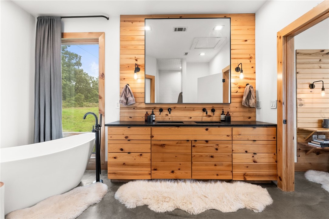 11334 Pool Holw Road Dardanelle, AR 72834 - Photo 20 of 50 Modern bathroom featuring a freestanding soaking tub with floor-mounted faucet, a double vanity with wood-finish cabinetry, black countertop, wall-mounted faucets, and a large framed mirror, all set against a backdrop of wood-paneled walls and concrete flooring