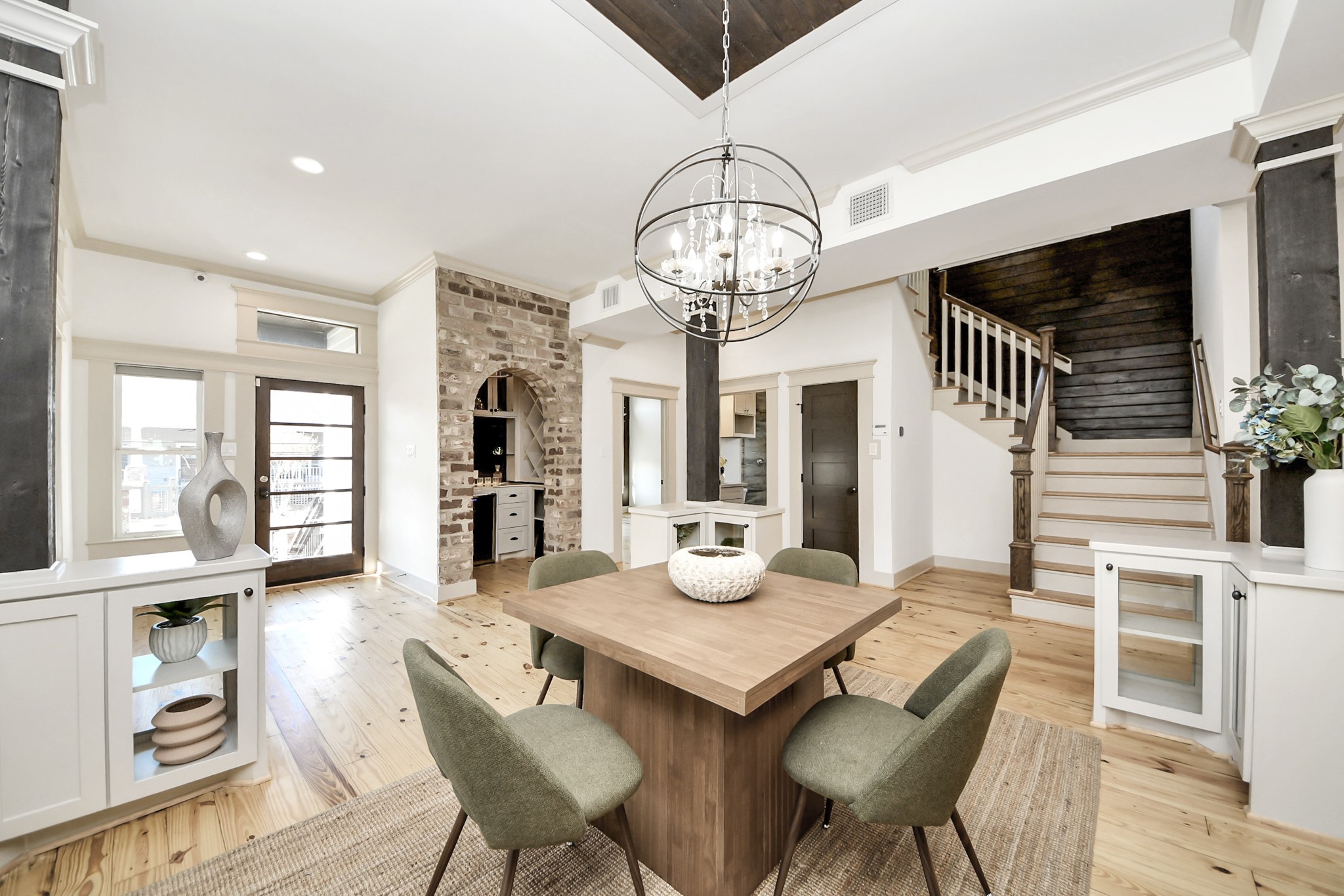 1202 Archer Street Houston, TX 77009 - Photo 16 of 46 This modern dining area features a sleek wooden table paired with stylish chairs, set against light hardwood floors that brighten the space. A character-rich brick accent wall, unique chandelier, and built-in shelving add depth and sophistication. The nearby staircase with dark wooden steps, along with the glass-paneled front door, introduces an elegant touch that completes the room’s inviting charm.