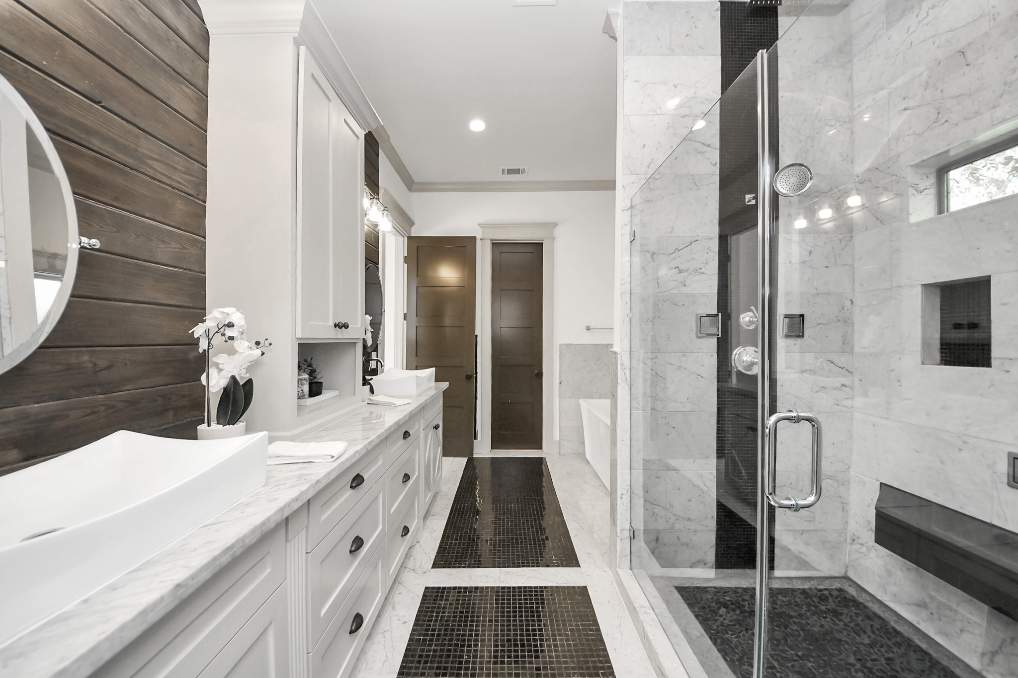 1202 Archer Street Houston, TX 77009 - Photo 24 of 46 This modern bathroom showcases a spacious glass-enclosed shower, a double vanity with sleek vessel sinks, and elegant marble countertops. A warm wood accent wall complements the neutral color palette, creating a clean, sophisticated, and inviting space.