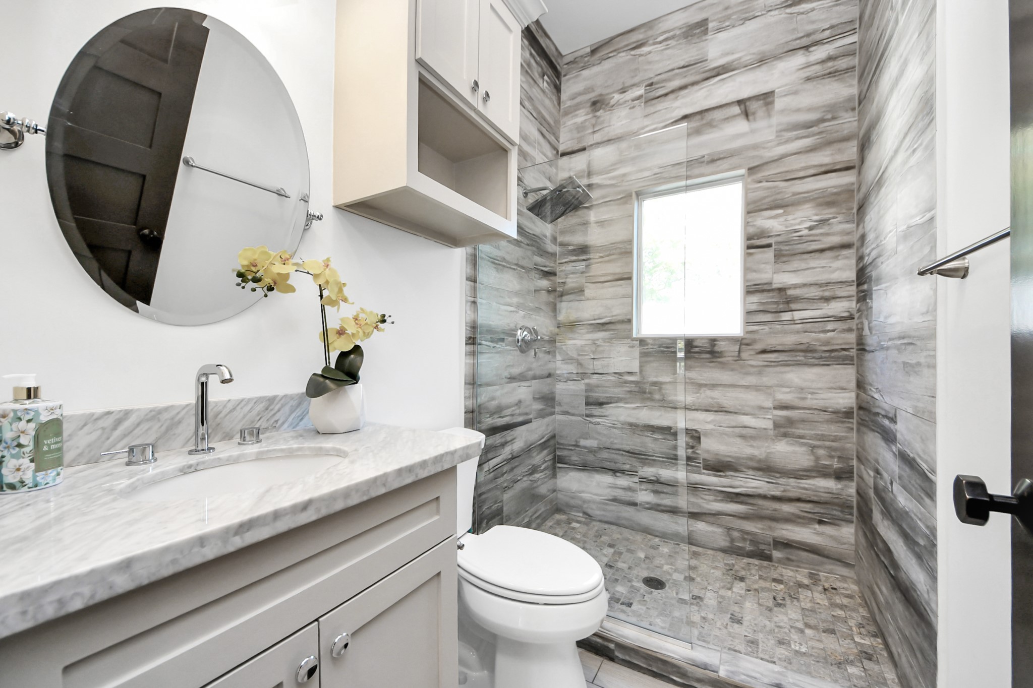 1202 Archer Street Houston, TX 77009 - Photo 32 of 46 This modern bathroom located on the ground level offers a sleek design with a round mirror, a marble countertop, and a frameless-enclosed shower. The shower area has stylish wood-look tiles and a small window providing natural light.