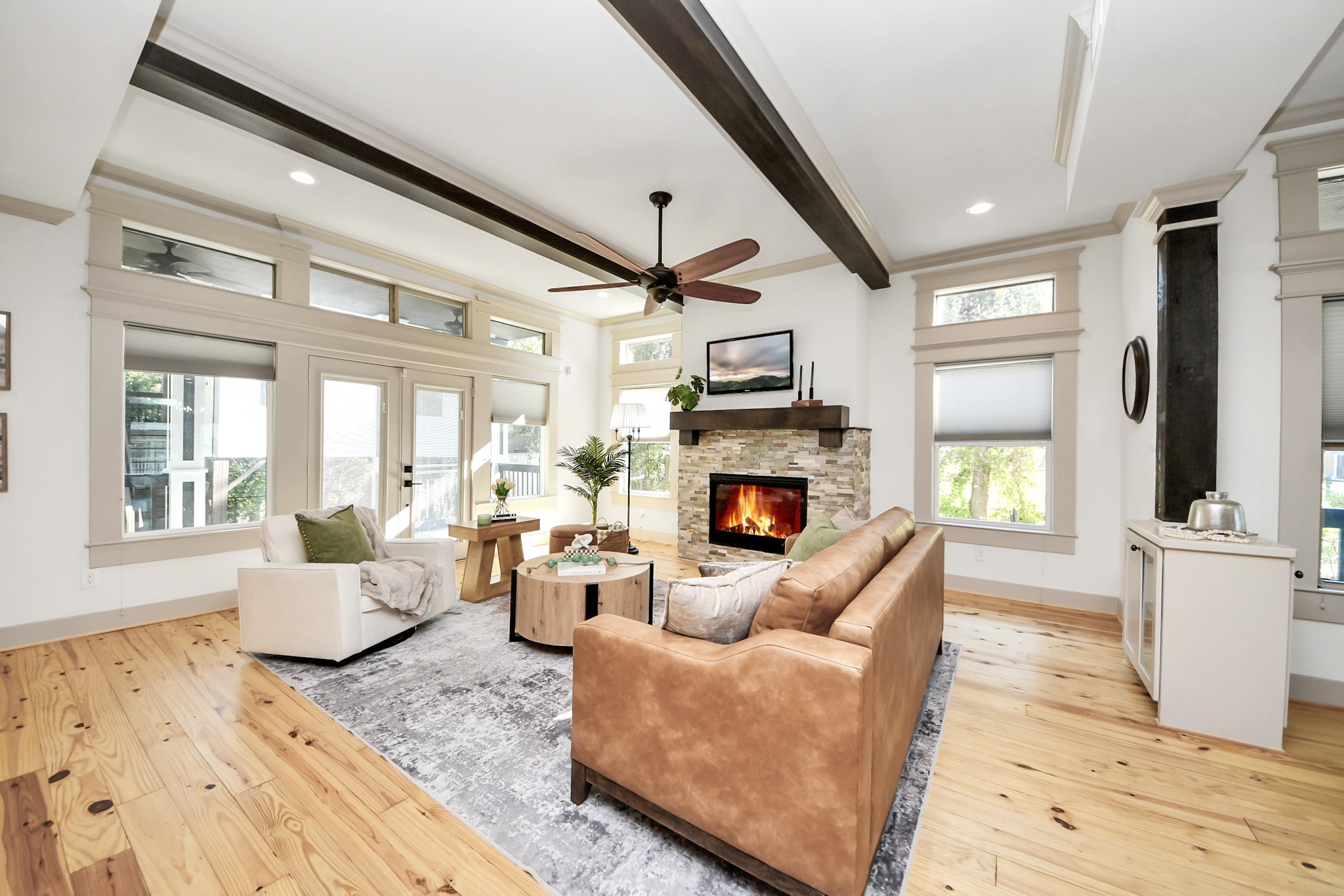 1202 Archer Street Houston, TX 77009 - Photo 8 of 46 This bright living room features a cozy stone fireplace, large windows for natural light, and exposed wooden beams. It has a comfortable seating area with a modern rug and a ceiling fan, creating a welcoming and airy atmosphere.