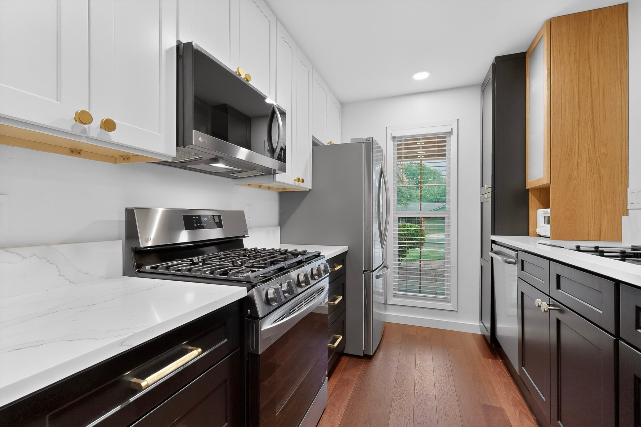 5541 Pine Place Austin, TX 78744 - Photo 12 of 33 Modern kitchen featuring white upper cabinetry and dark lower cabinetry with gold-toned hardware, stainless steel appliances, white countertops, and wood-finish flooring