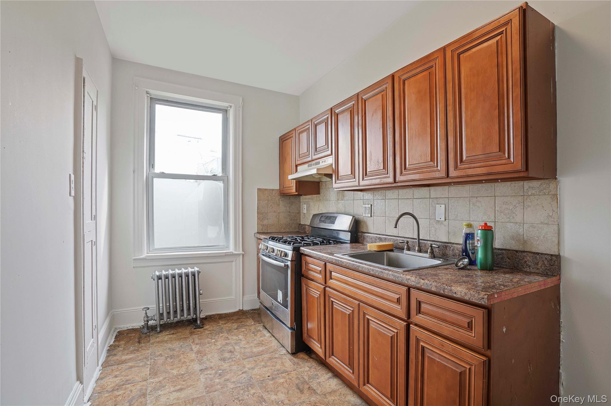 72-07 73rd Street Queens, NY 11385 - Photo 25 of 44 Kitchen featuring dark countertops, stainless steel gas stove, brown cabinetry, radiator heating unit, and tasteful backsplash