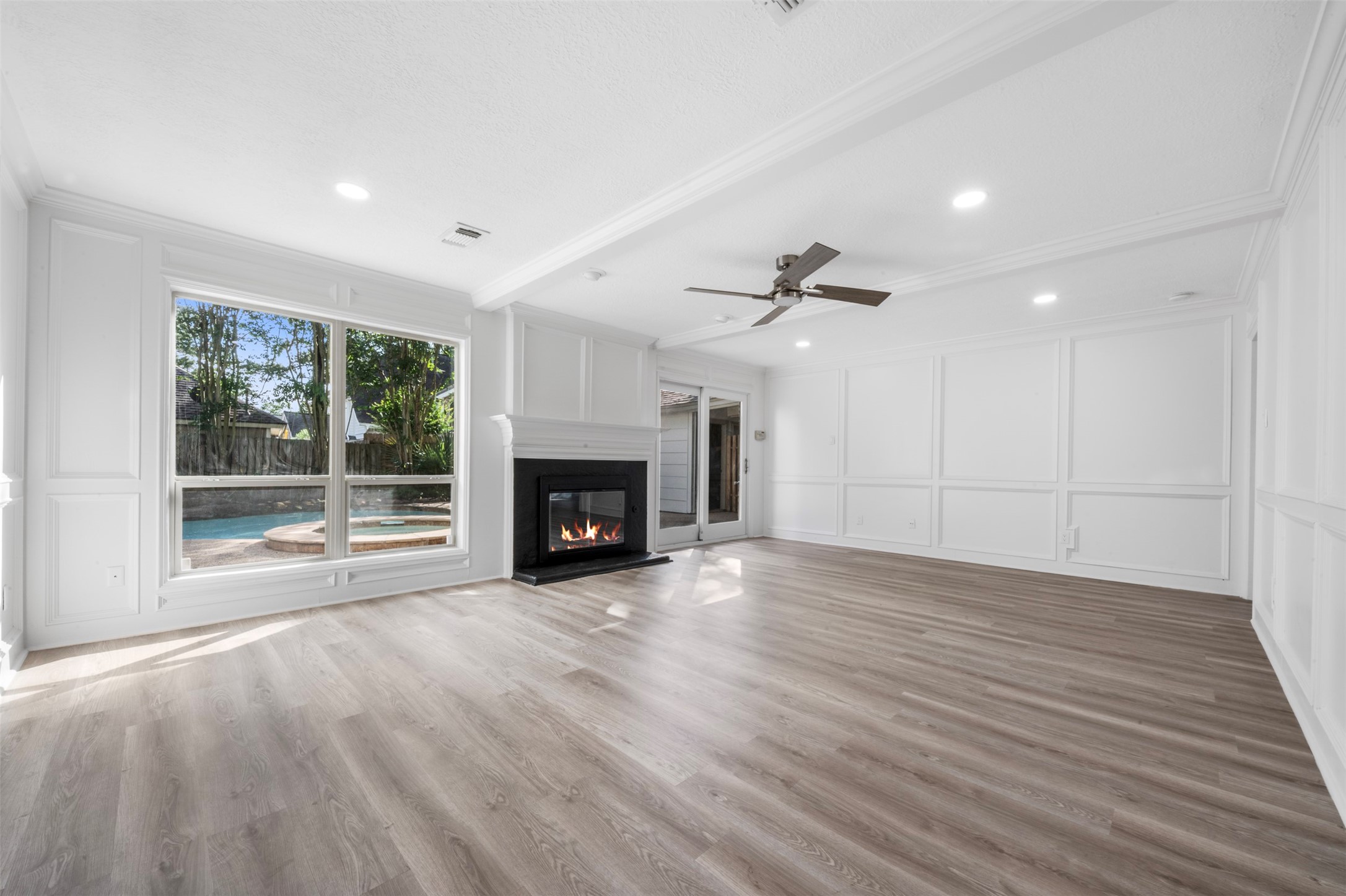 9543 Enstone Circle Spring, TX 77379 - Photo 3 of 50 Bright and inviting living room with large windows offering a view of the backyard pool. Features include a modern fireplace, ceiling fan, and elegant wainscoting, complemented by light wood flooring.